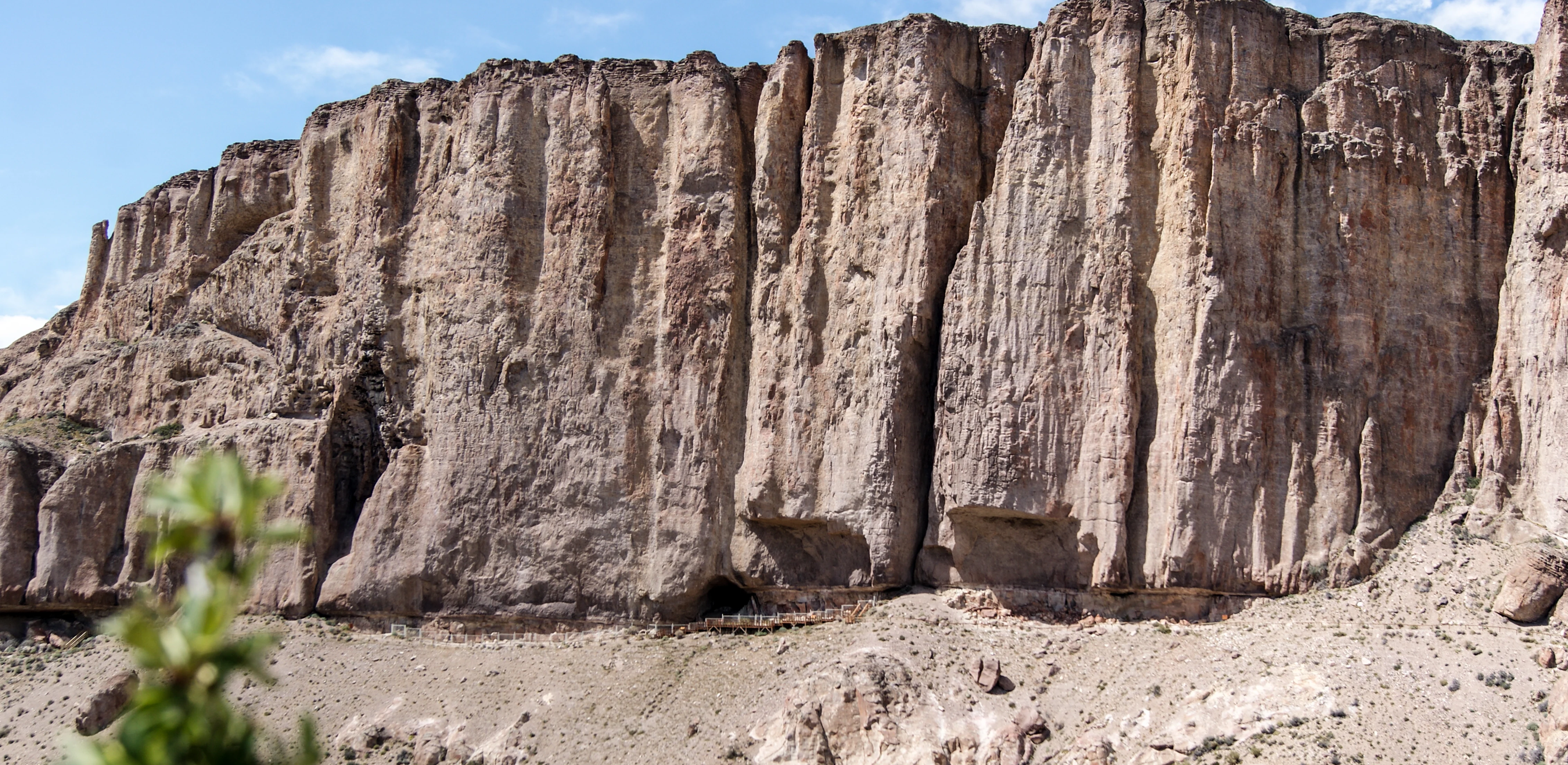 A wide view of a tall, vertical rock canyon wall with distinct vertical striations and a dark cave opening near the base.