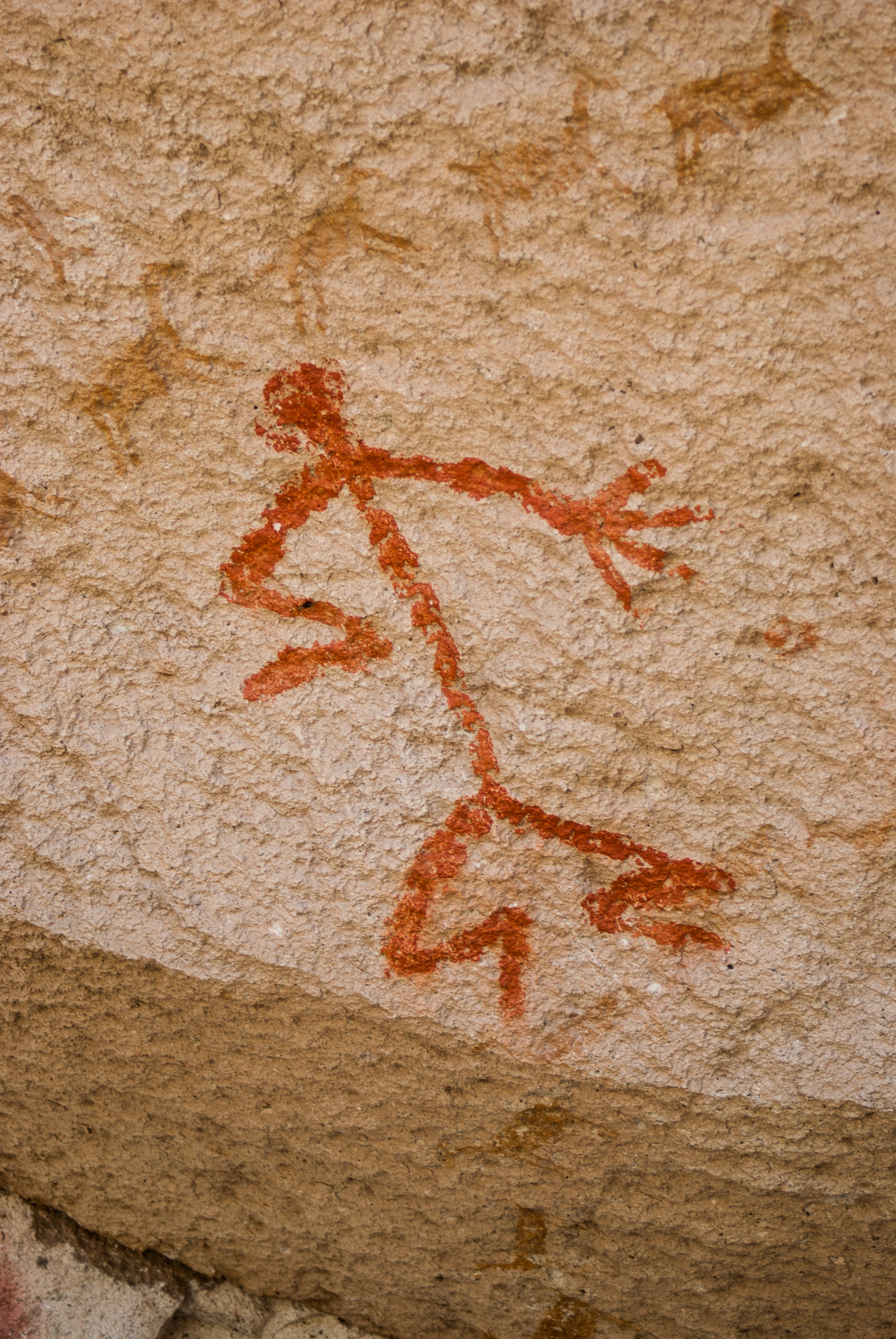 A close-up view of a prehistoric red pigment painting on a textured, light-colored rock surface, depicting a stylized human figure.