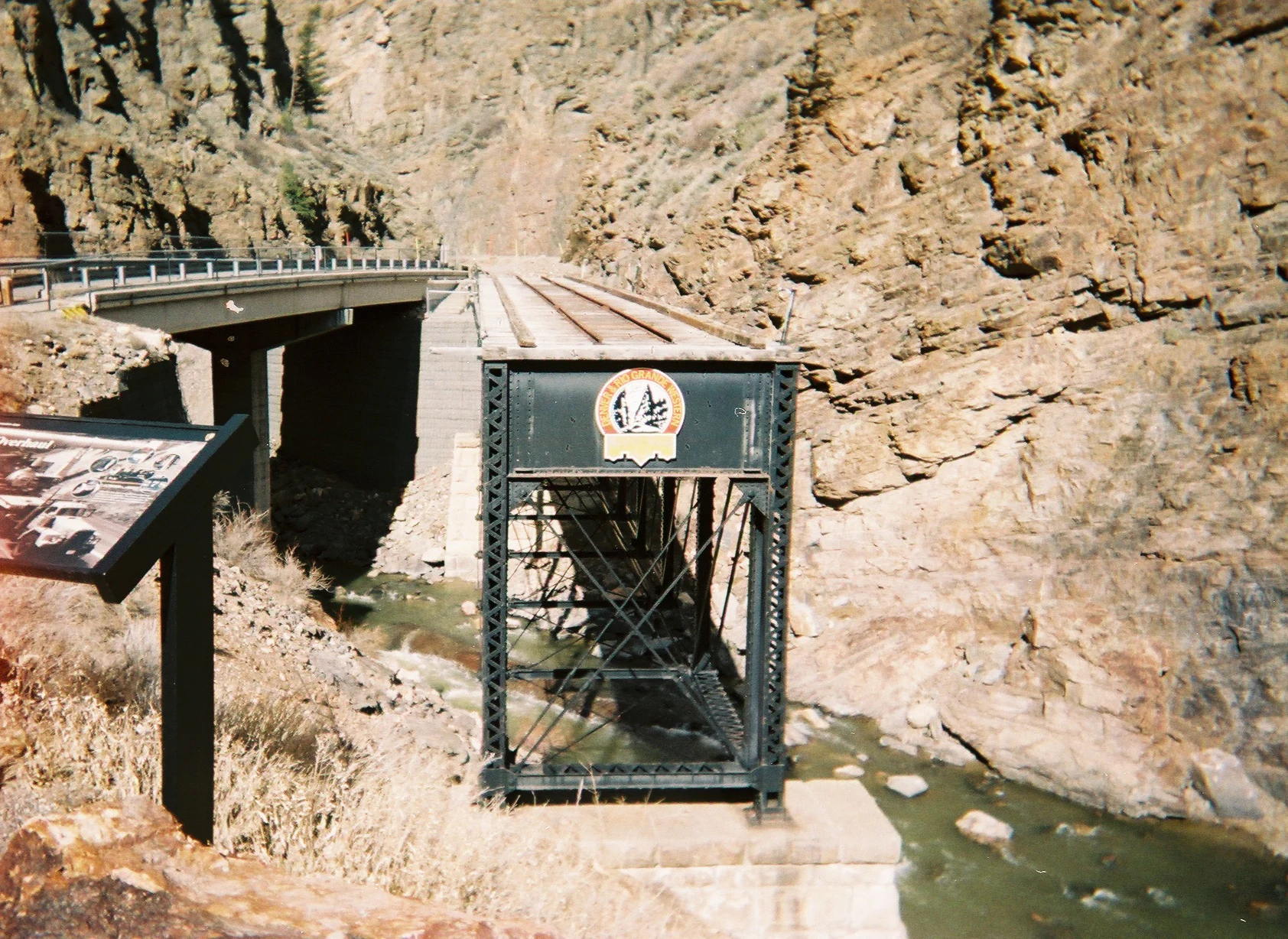 A vintage metal railroad bridge preserved on stone abutments, positioned near a modern highway bridge in a rocky canyon with a stream below.