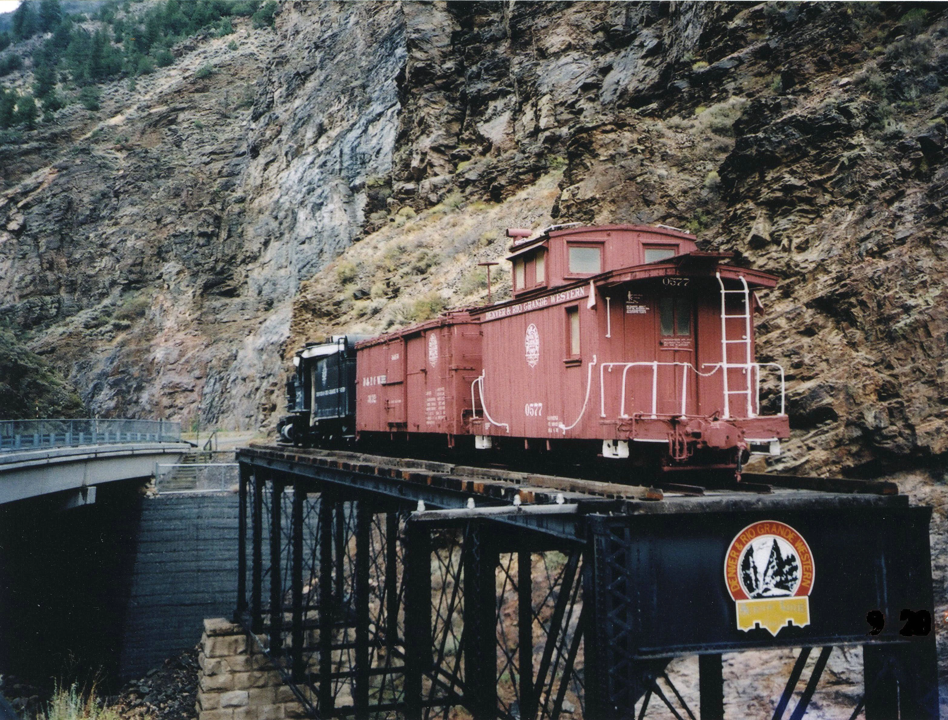 A red historic narrow gauge caboose sits on a short section of railway track on a steel trestle bridge in front of a steep rocky canyon wall.