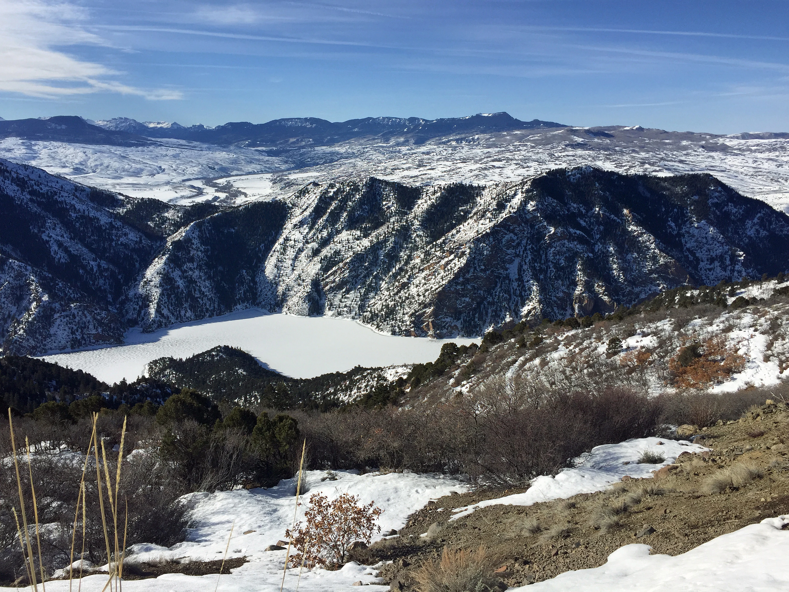 A wide, elevated view of a frozen, white, snow-covered reservoir nestled within a deep canyon; rugged mountains covered in patchy snow rise in the background under a clear blue sky.
