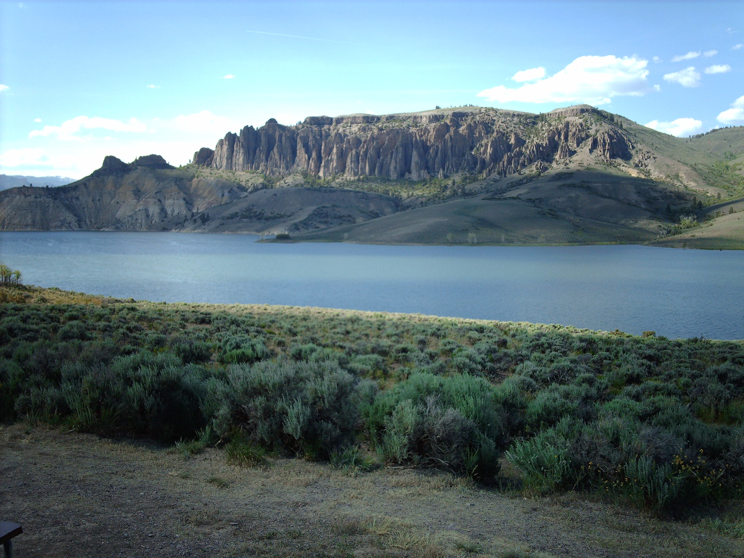 A wide scenic view of the Dillon Pinnacles, a prominent rock formation with vertical spires, set against a large reservoir and a clear blue sky, with sagebrush in the foreground.