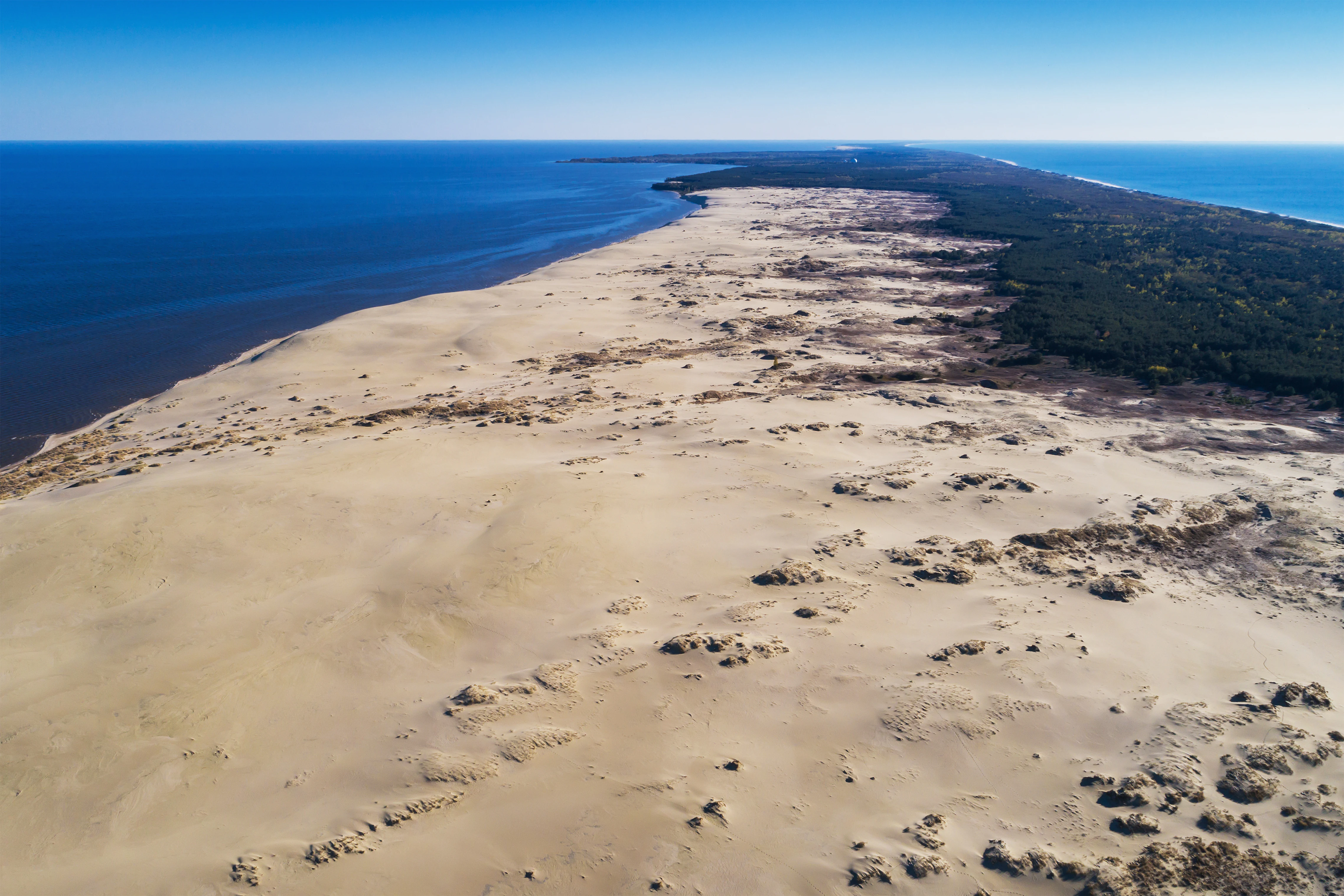 Aerial view of a sandy coastal spit extending into the sea, bordered by forested land and blue water, with visible sand dunes and sparse vegetation