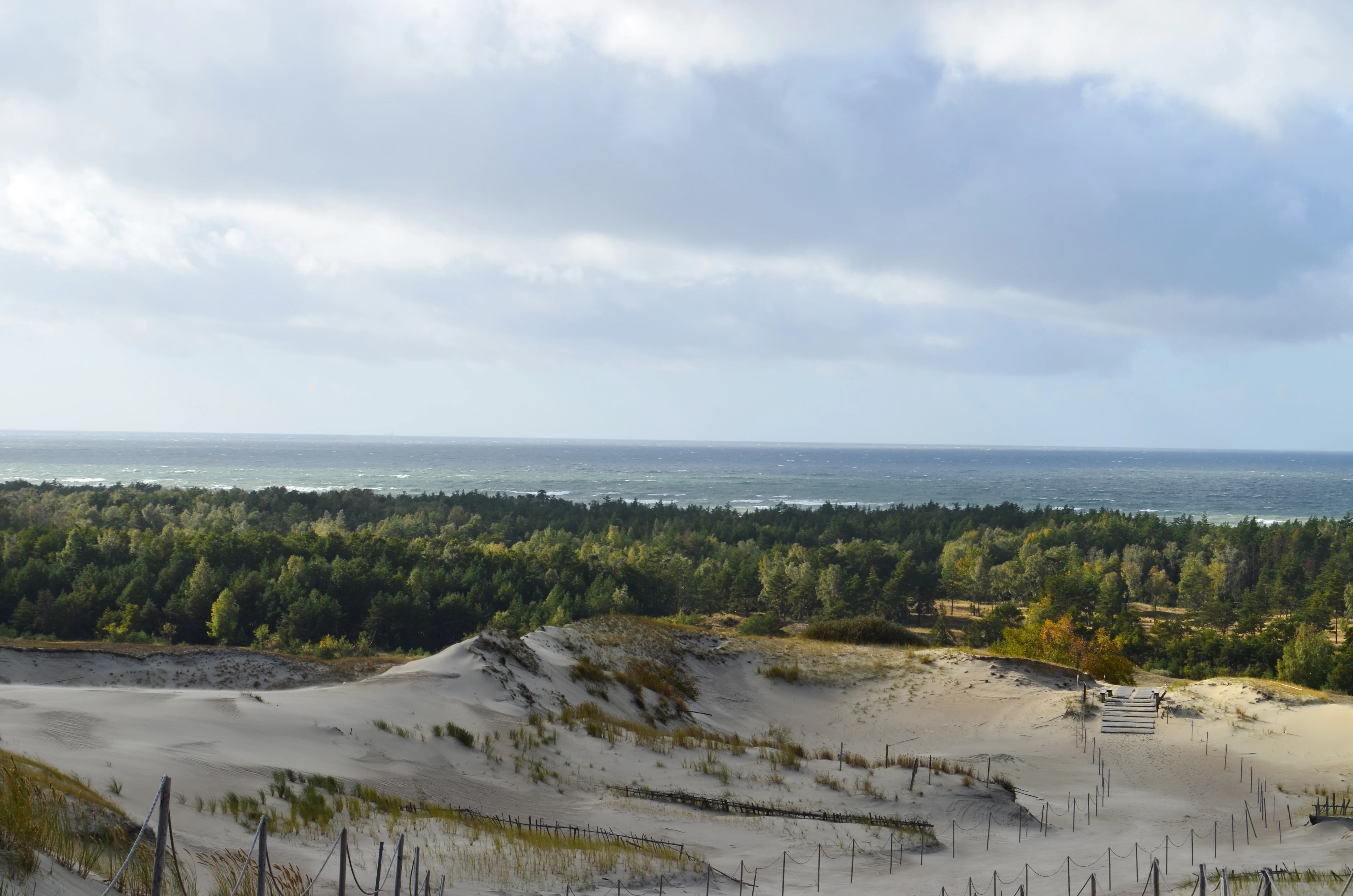 Sand dunes with sparse vegetation, a forested area in the midground, and the Baltic Sea under a partly cloudy sky