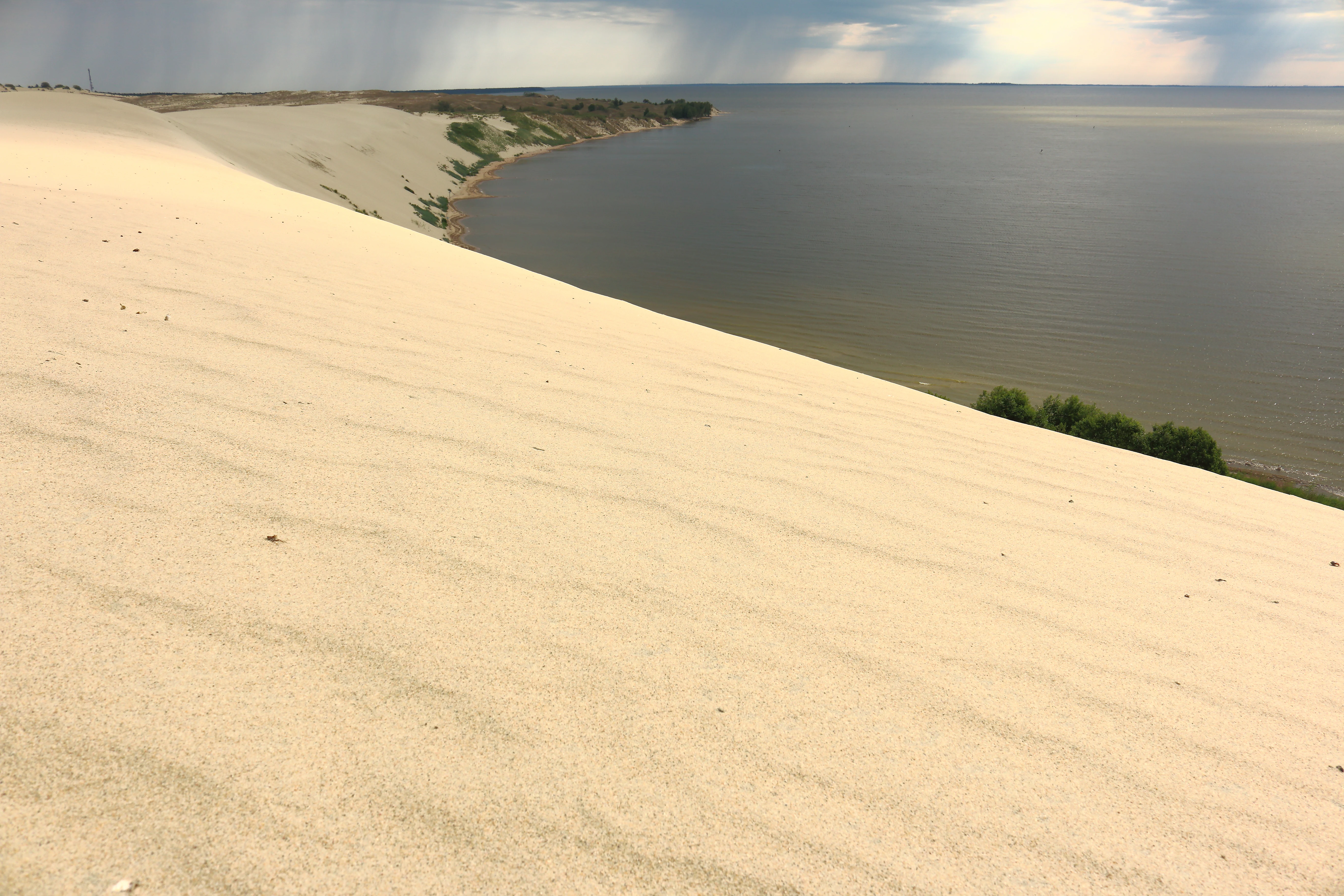 Large sand dune with wind-swept ridges slopes toward calm Curonian Lagoon water with distant shoreline vegetation