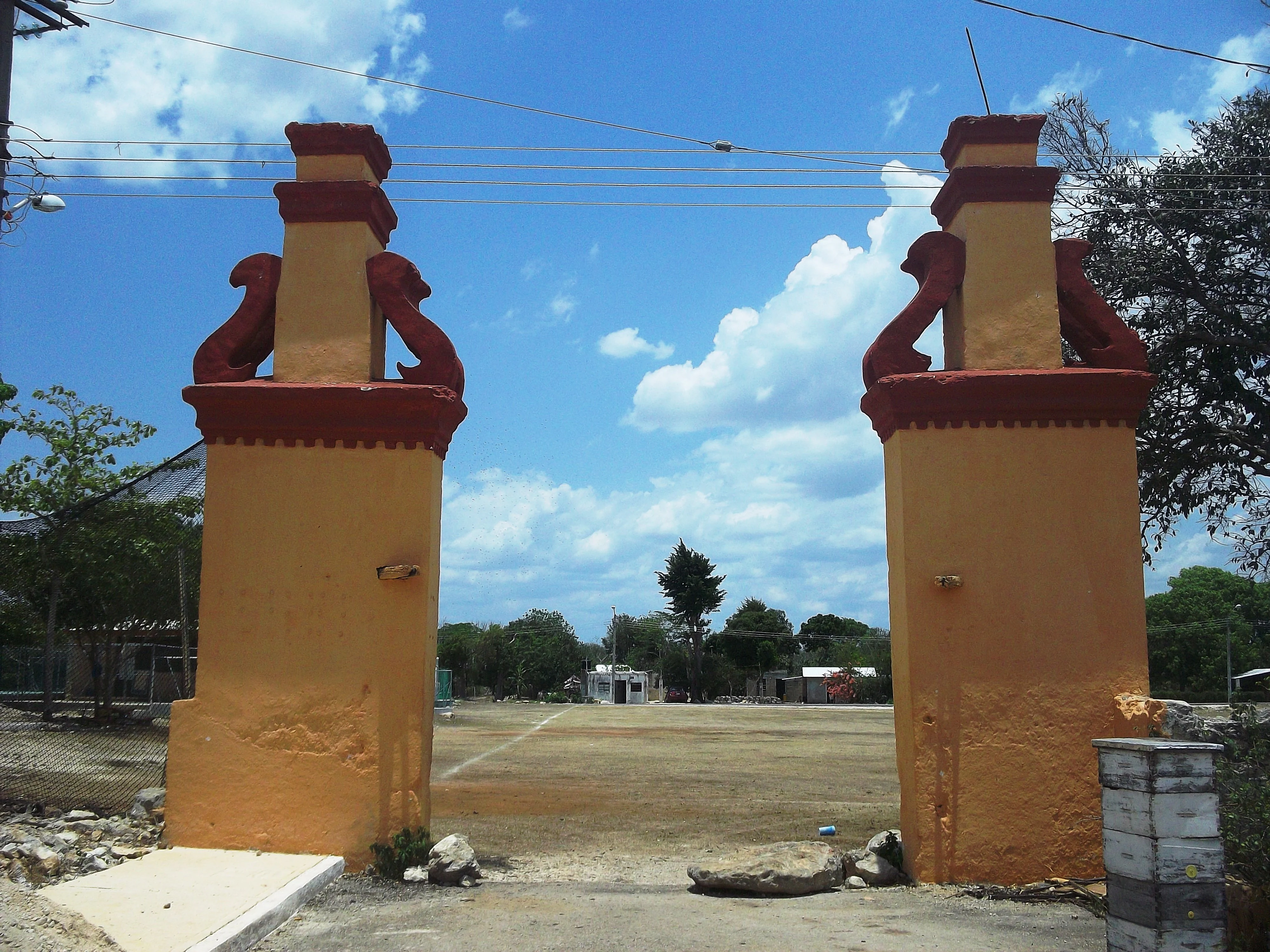 Two tall yellow and red stucco pillars forming a gate, leading to an open dirt field under a bright blue sky.