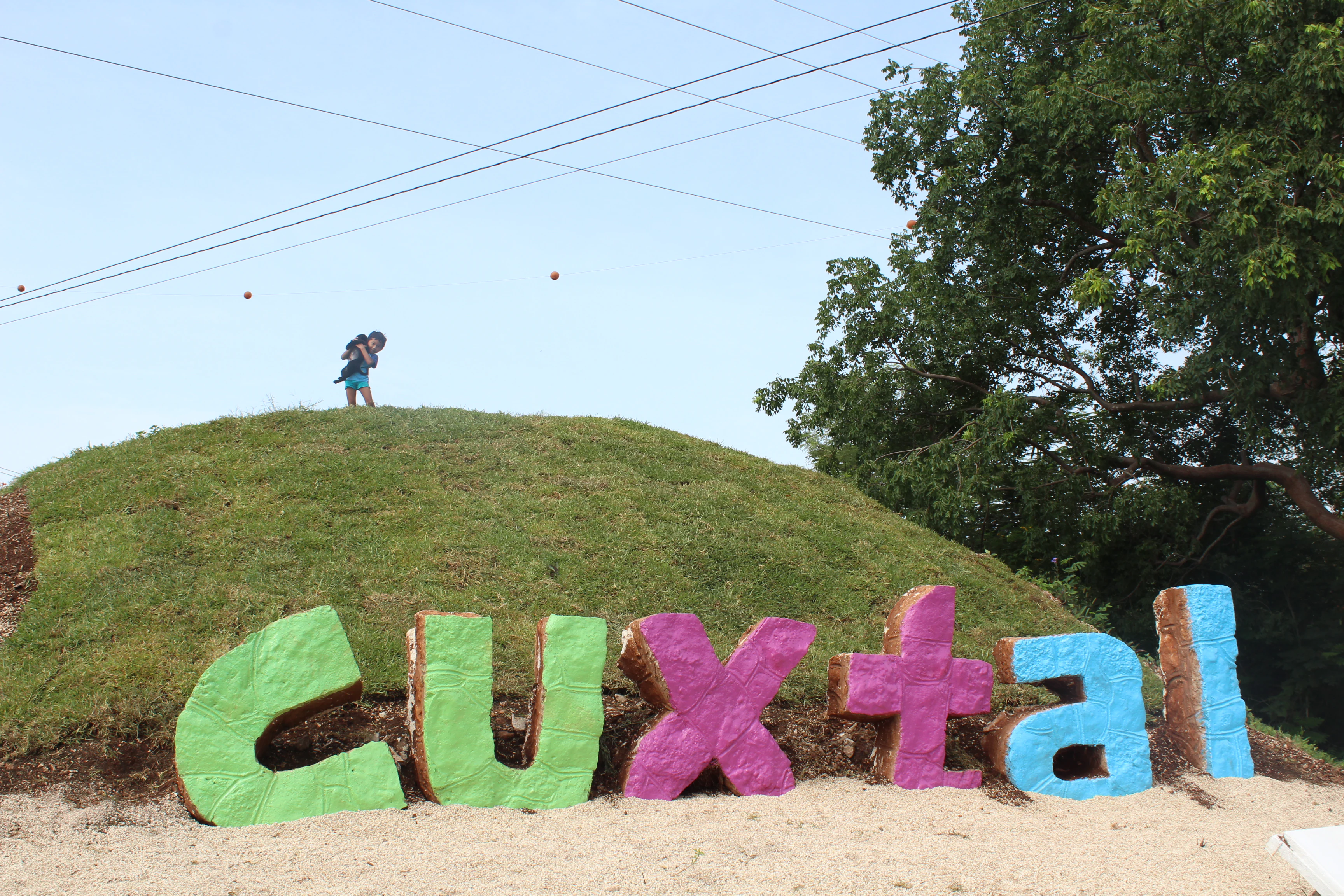 A large, multi-colored sign reading Cuxtal placed on a grassy hill, with a person standing on top of the hill.