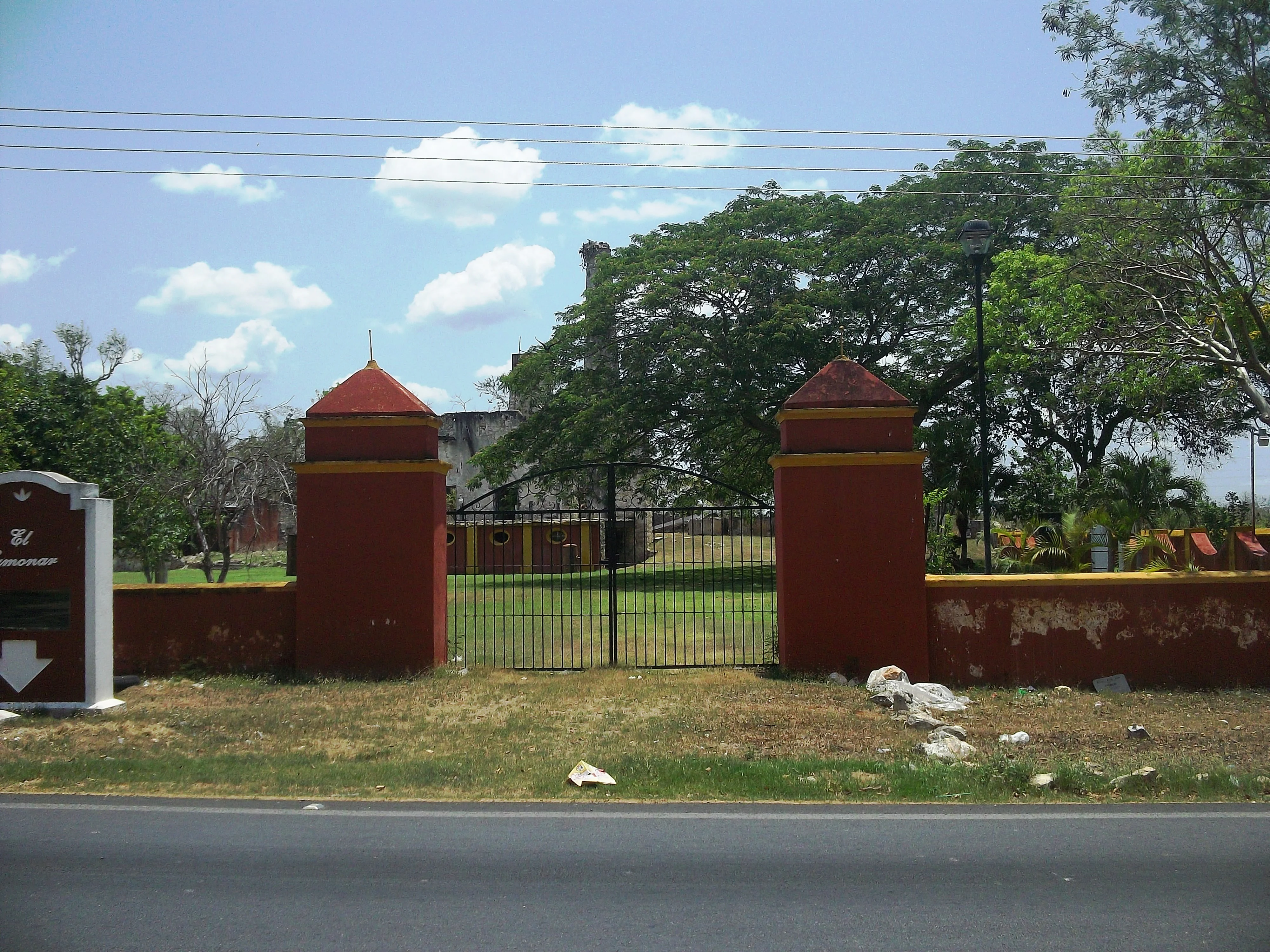 A red and yellow stucco gate and wall with a black metal fence, marking the entrance to a hacienda property with trees in the background.