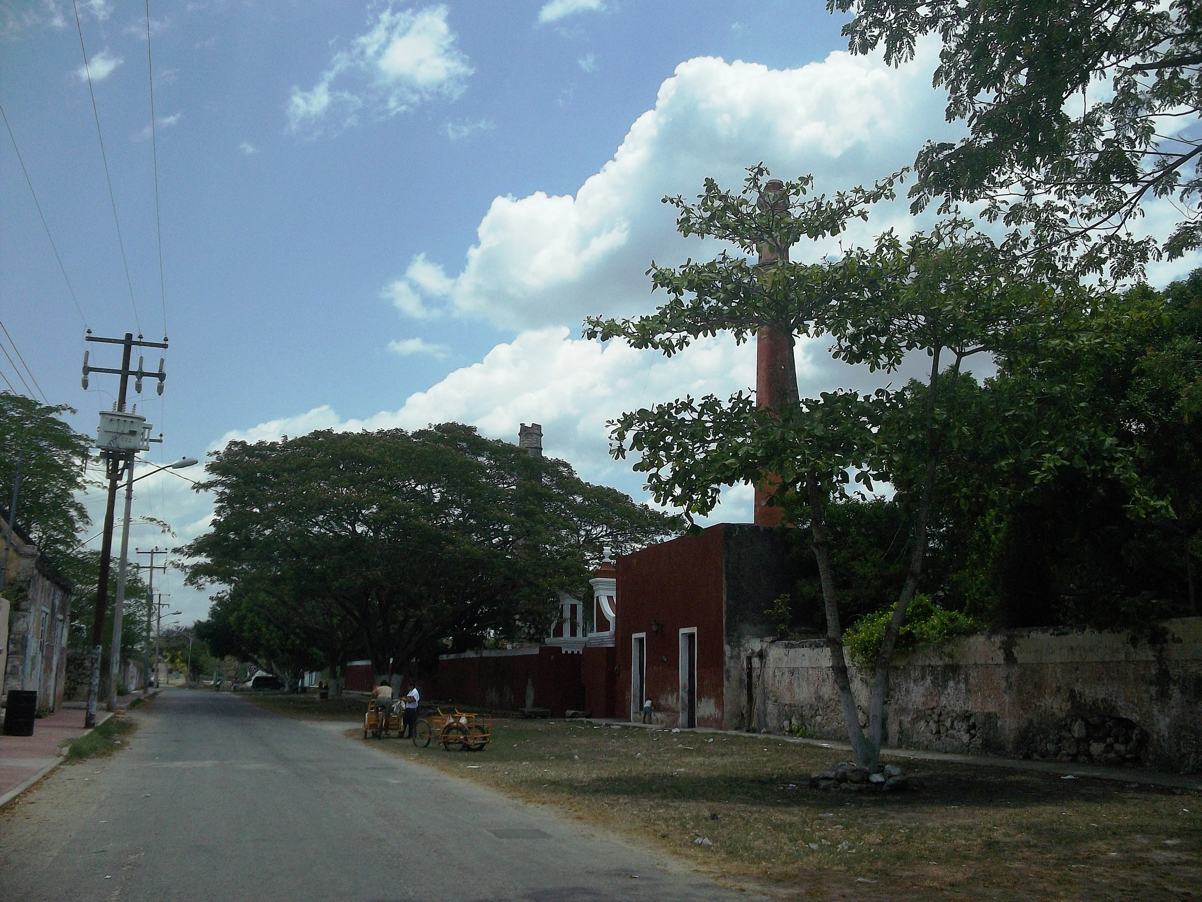 A paved road leading past the exterior walls and trees of the historic Hacienda San Pedro Chimay under a blue sky with white clouds.