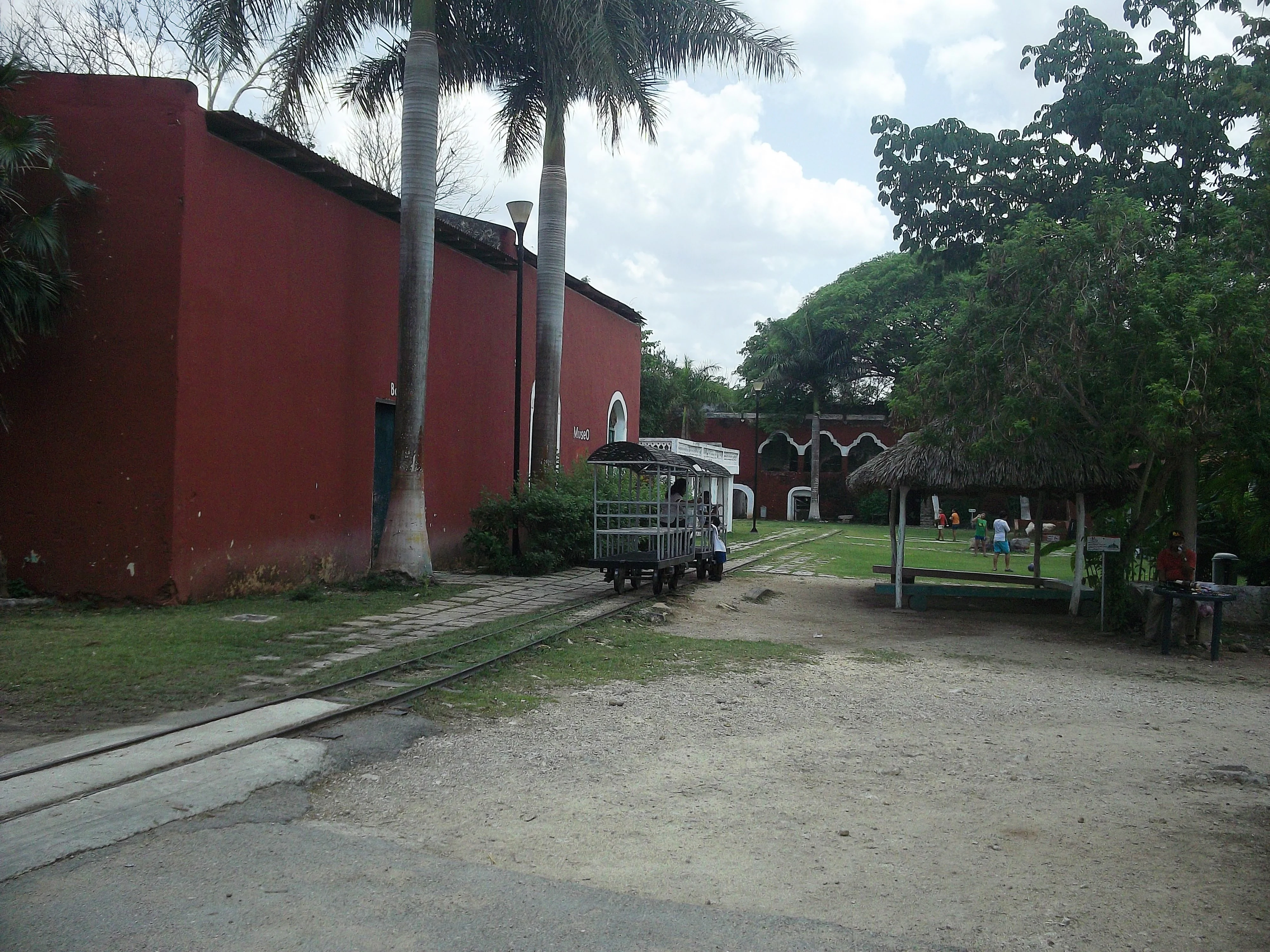 A red colonial-style building and palm trees next to a small tourist train on tracks in the Cuxtal Ecological Reserve.