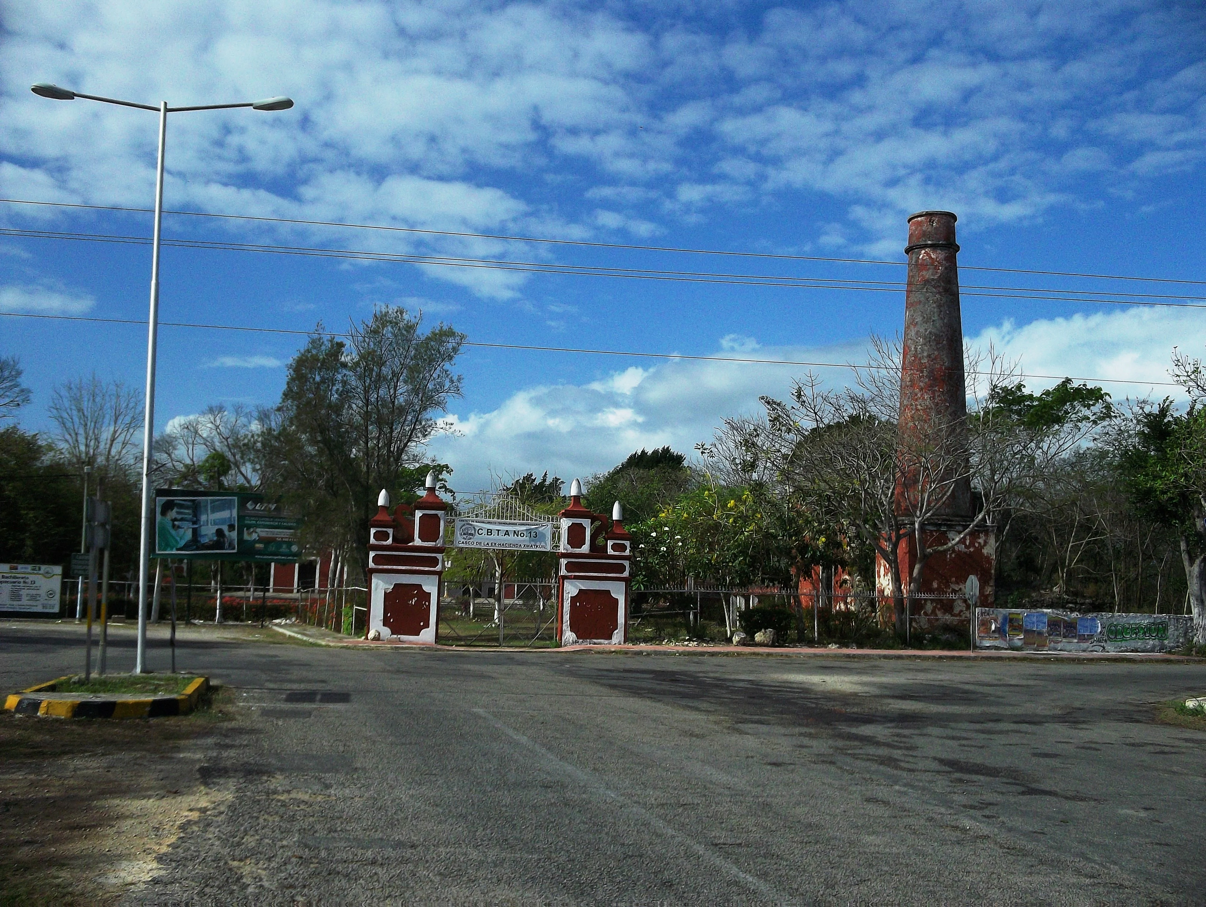An asphalt road leads toward a gated entrance with white and red masonry columns, featuring a tall, slender brick chimney standing on the right side under a blue sky.