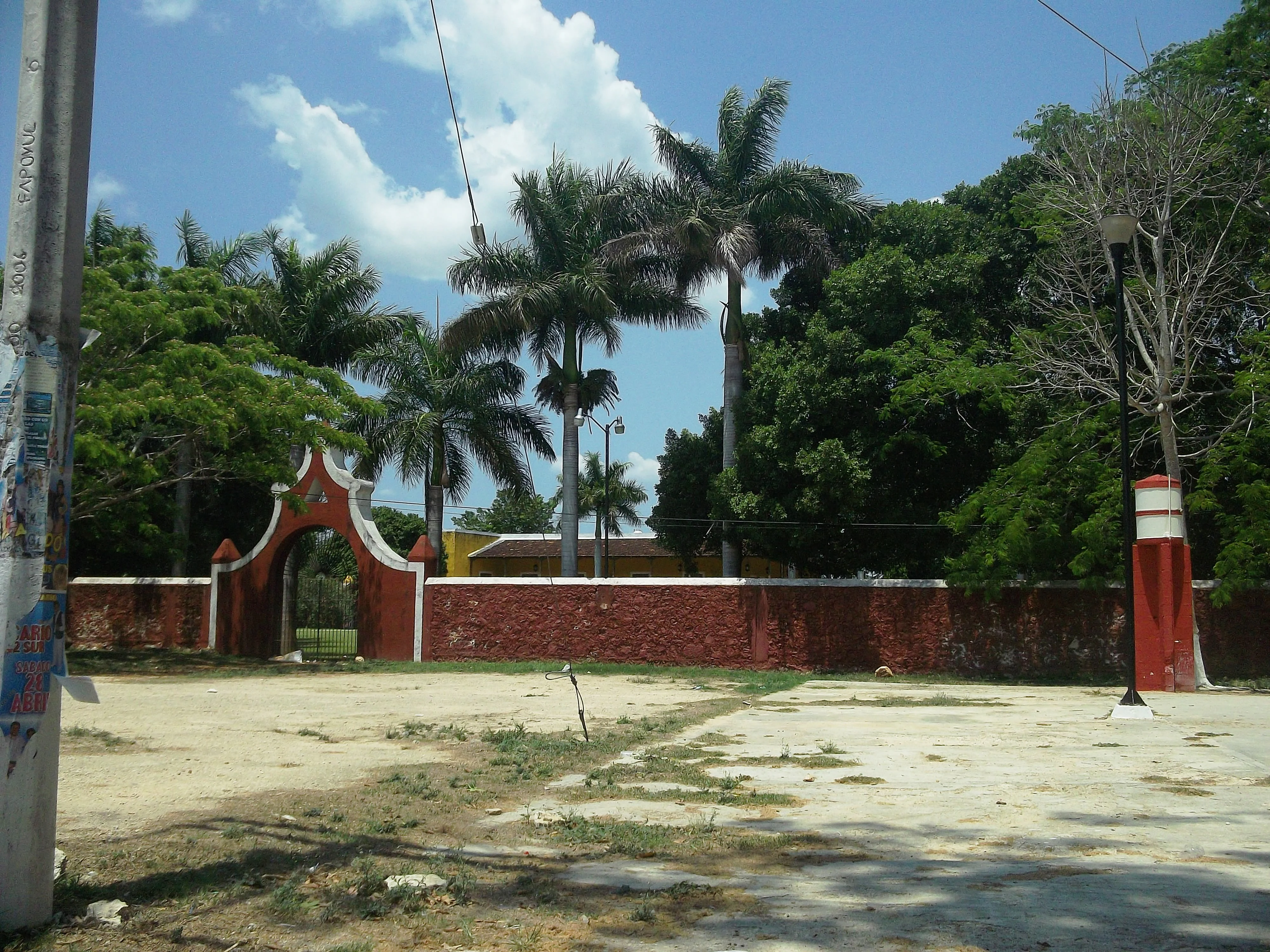 A red brick wall with an arched entrance gate set against a backdrop of palm trees and other lush greenery under a clear sky.