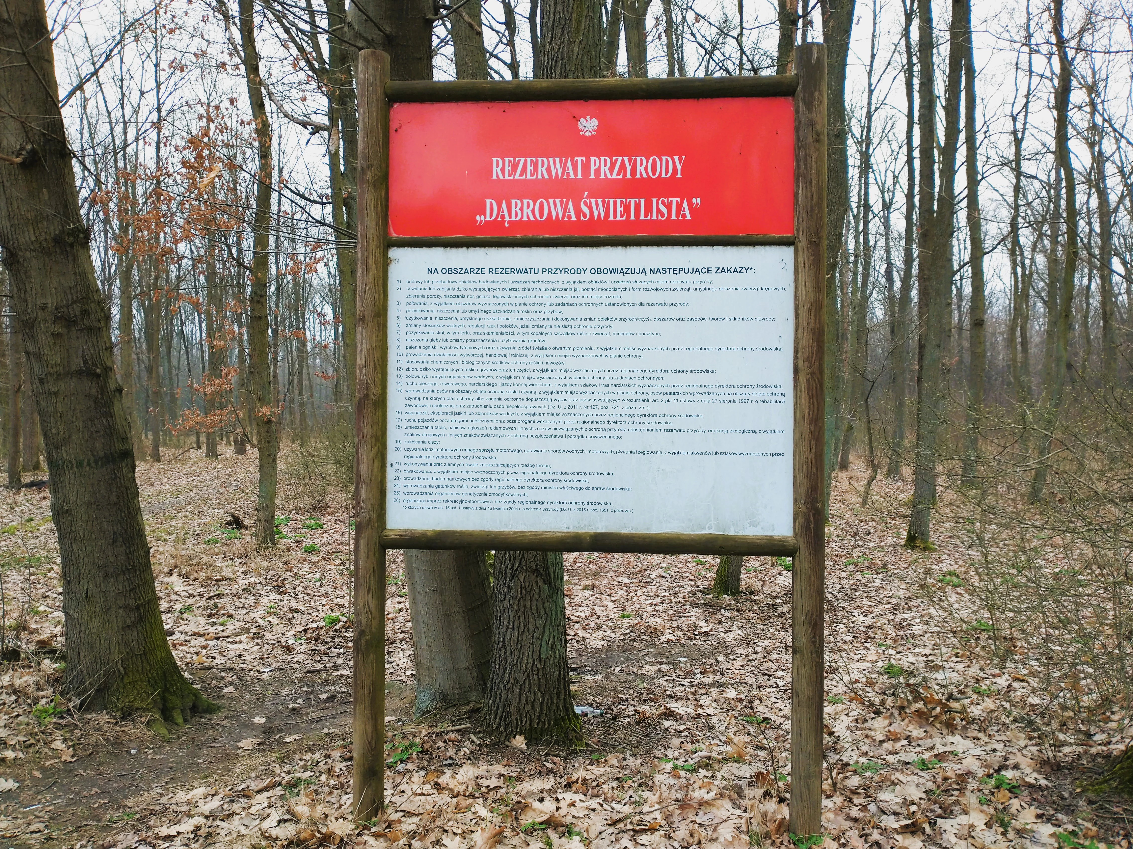 A rectangular information sign standing on two wooden posts in a forest setting. The sign has a red header with the text REZERWAT PRZYRODY DĄBROWA ŚWIETLISTA and a large white lower section containing detailed regulations and park rules in Polish text.