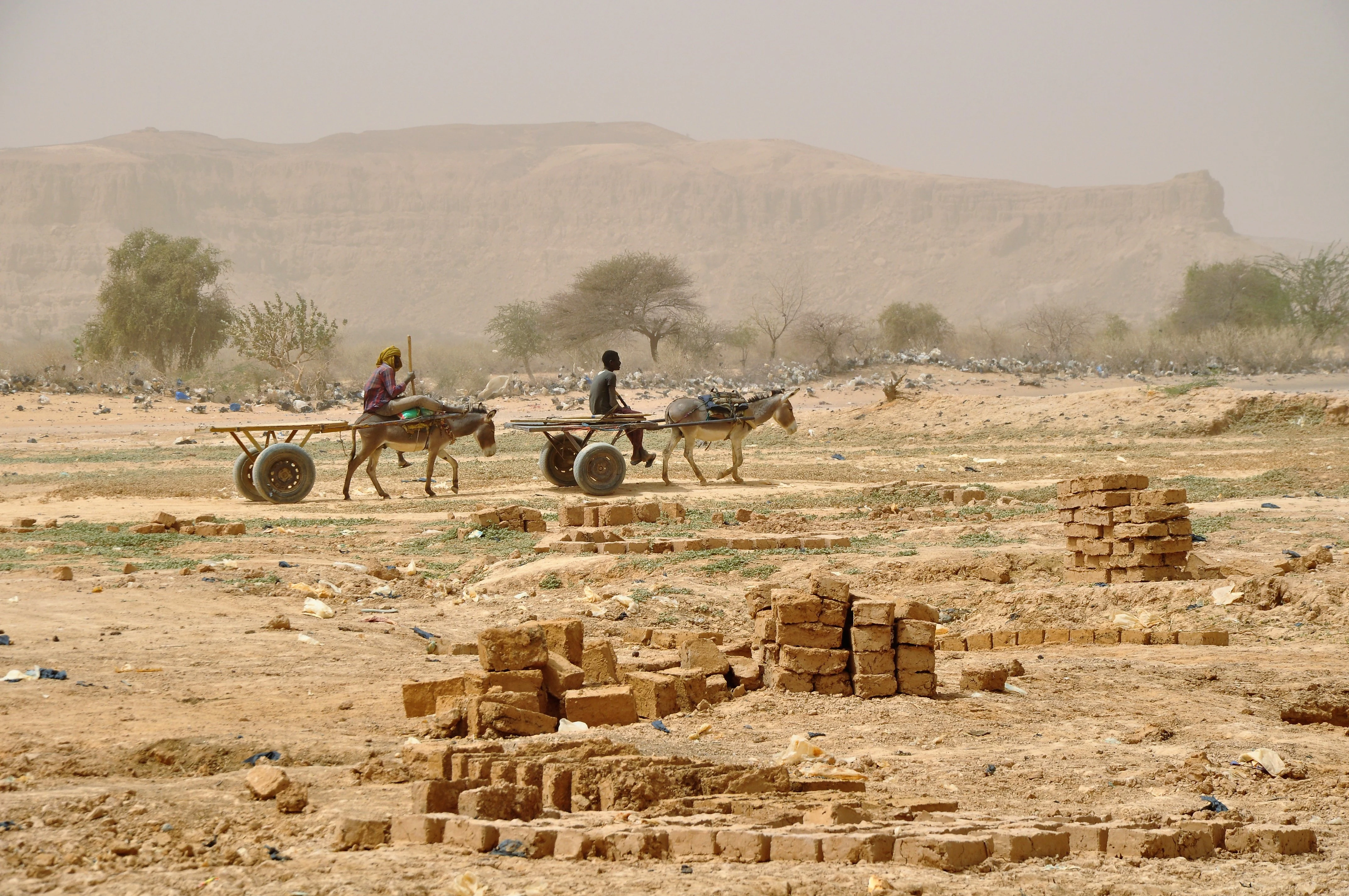 Two people riding in donkey-drawn carts across a dry, arid landscape with a large escarpment in the background and stacks of mud bricks in the foreground.