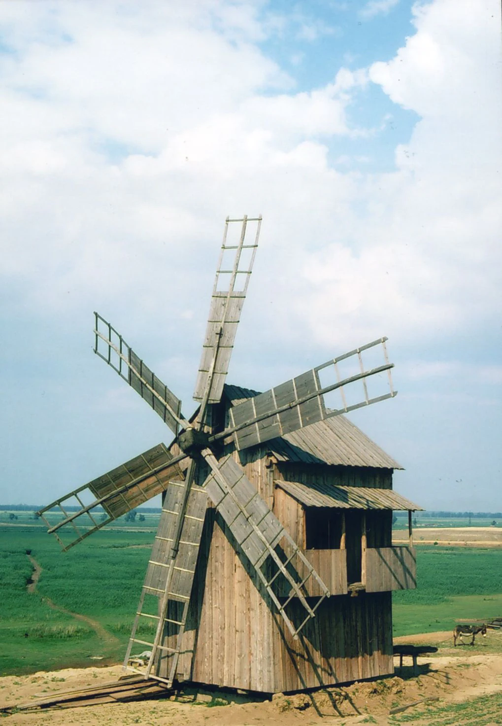 Wooden windmill with ladder-like blades in a grassy field under a partly cloudy sky