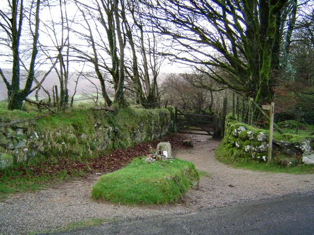 Stone grave marker on grassy mound surrounded by stone walls, trees, and gravel path with a gate in the background