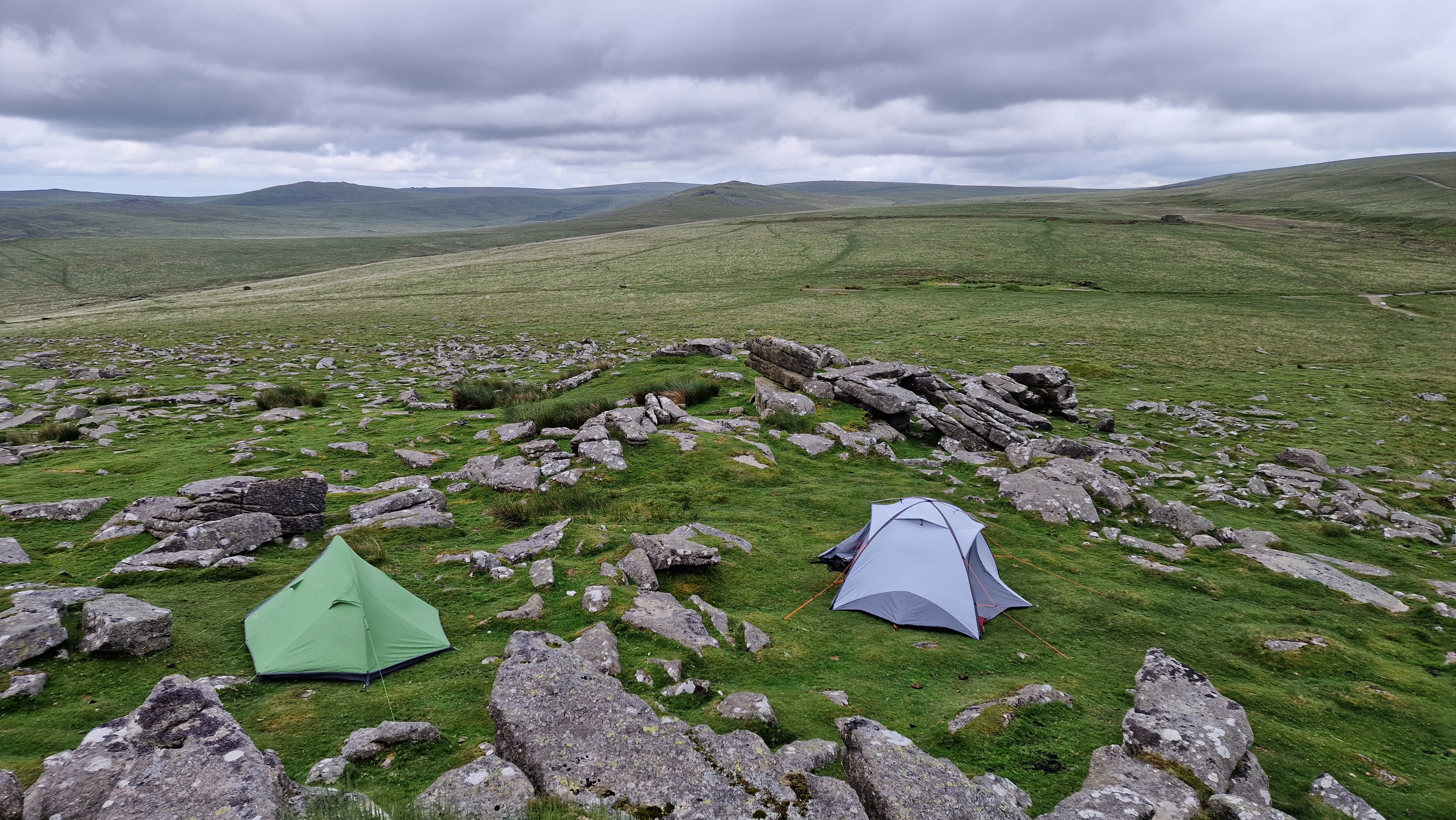 Green and gray tents set up on a rocky grassy moorland with rolling hills and a cloudy sky