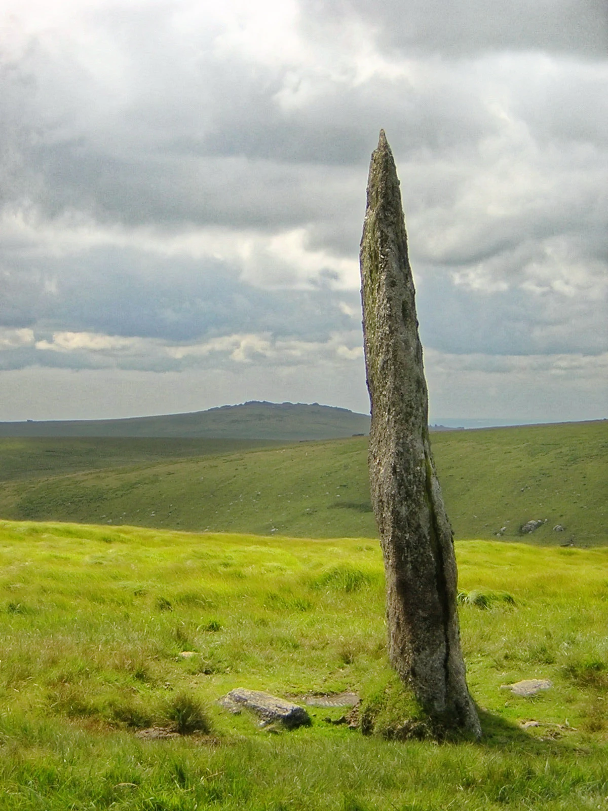 Tall weathered standing stone in a grassy moorland field with rolling hills under a cloudy sky