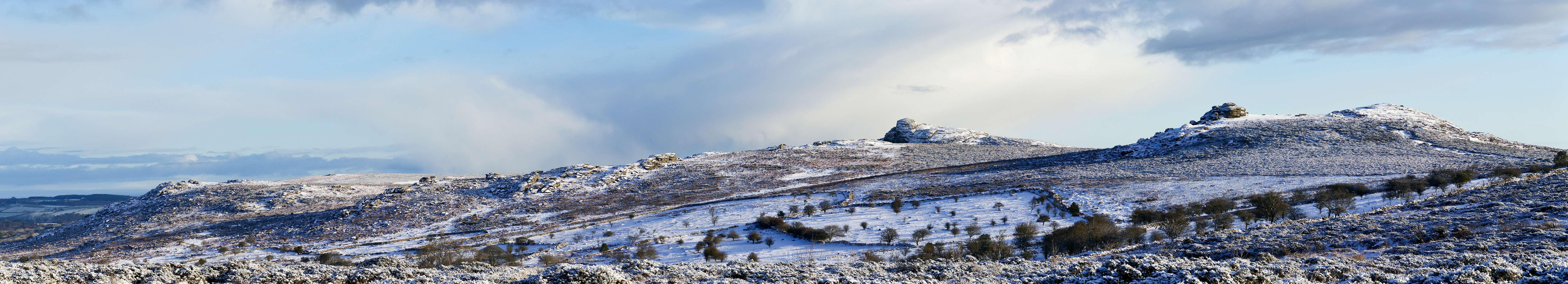 Wide panoramic view of snow-covered granite tors on Dartmoor moorland under a partly cloudy sky