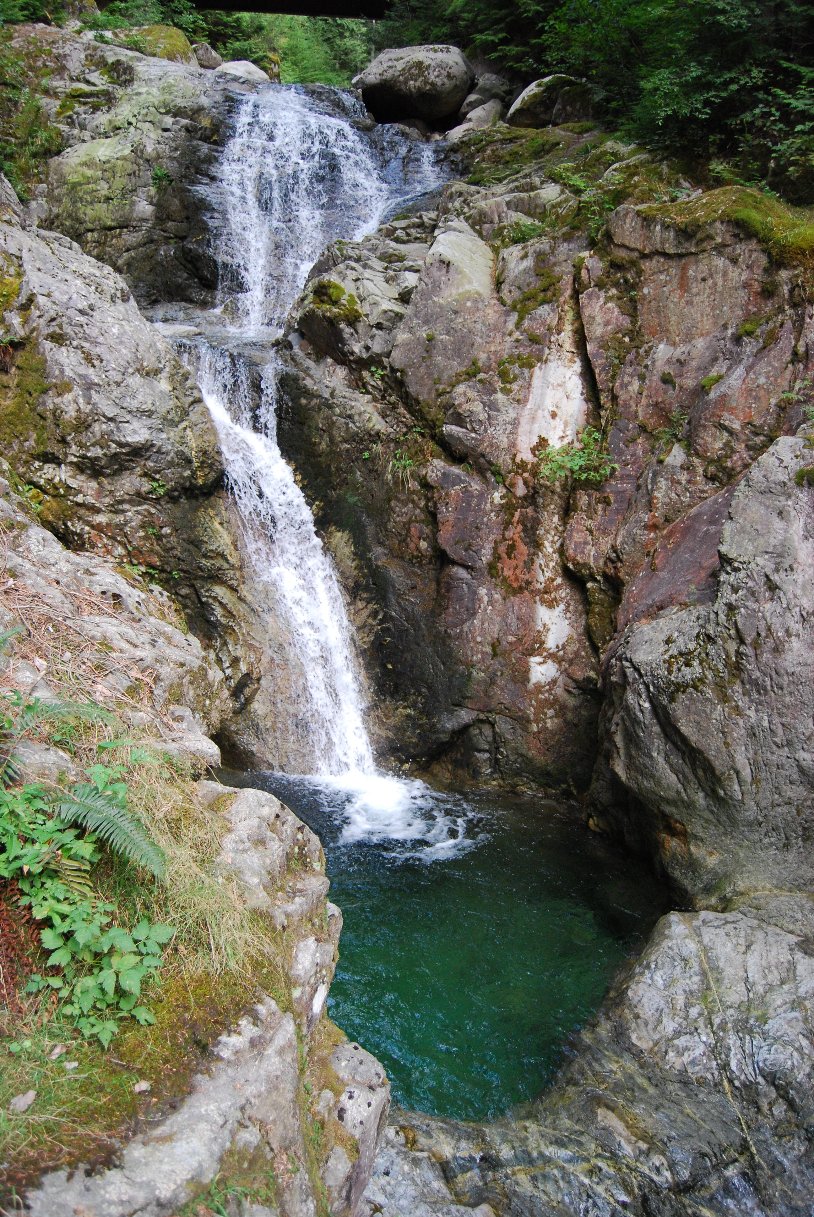 A multi-tiered waterfall cascading over dark rocky cliffs into a clear, deep green pool in a forested area.