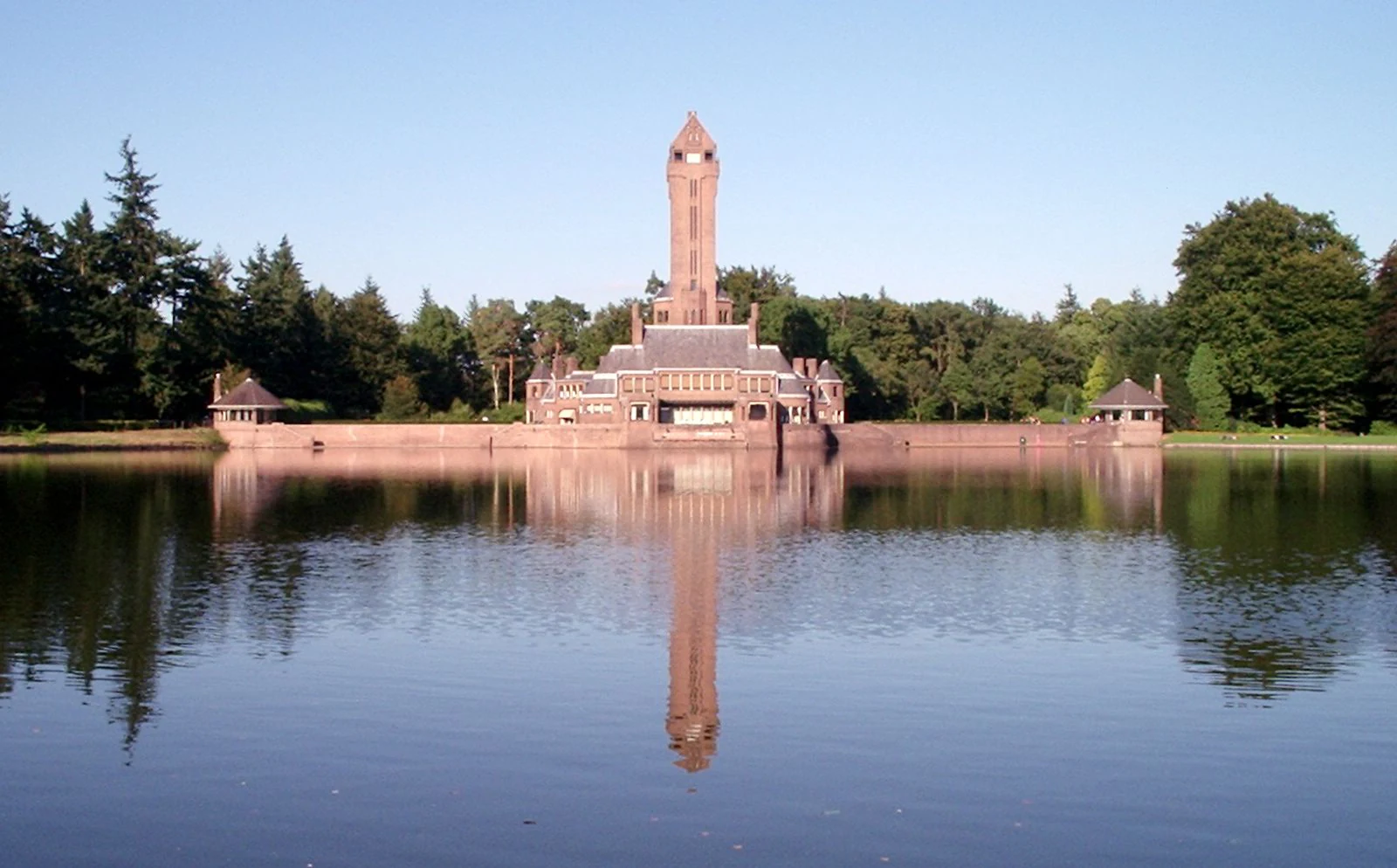 Large brick building with a tall tower reflected in a still lake surrounded by trees