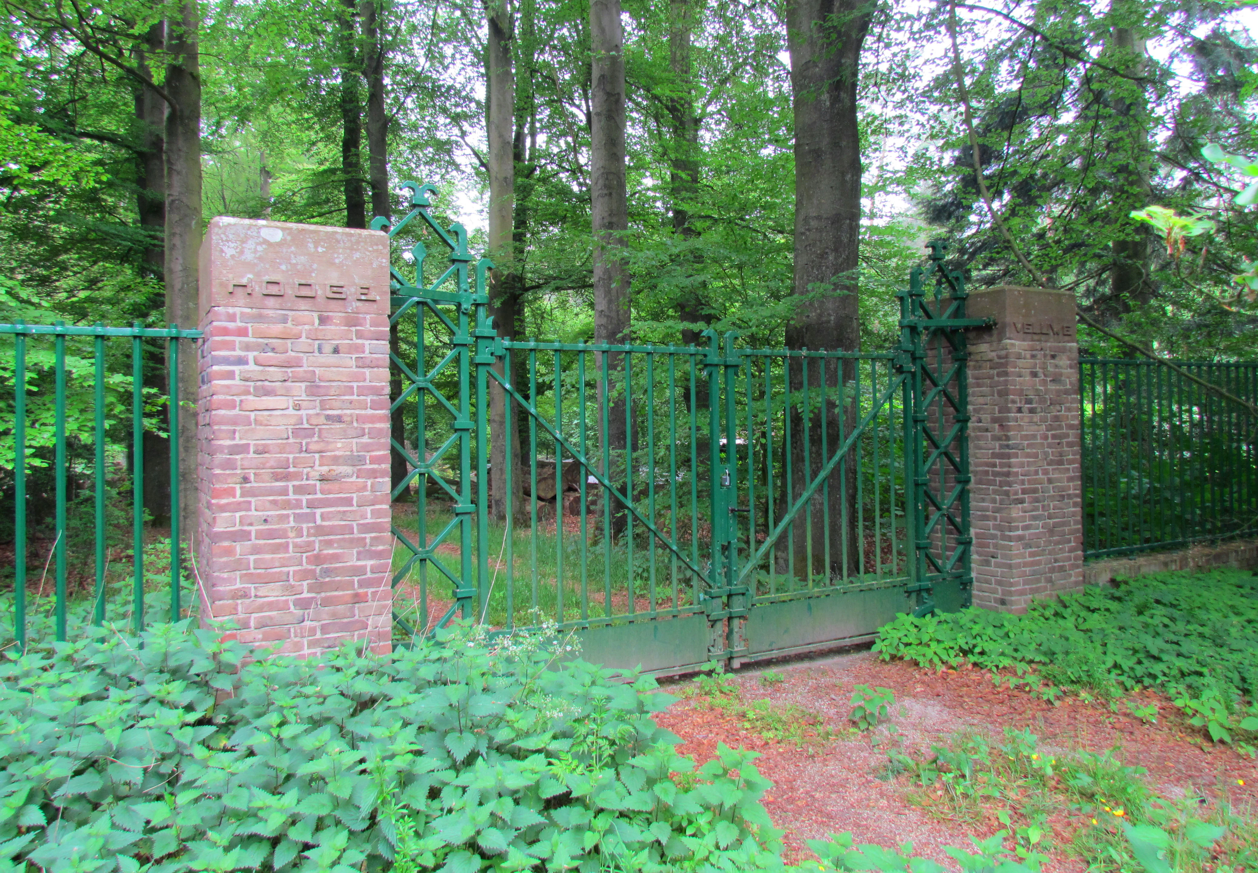 Green metal gate flanked by brick pillars with 'HOENDERLOO' text, surrounded by dense green foliage and trees