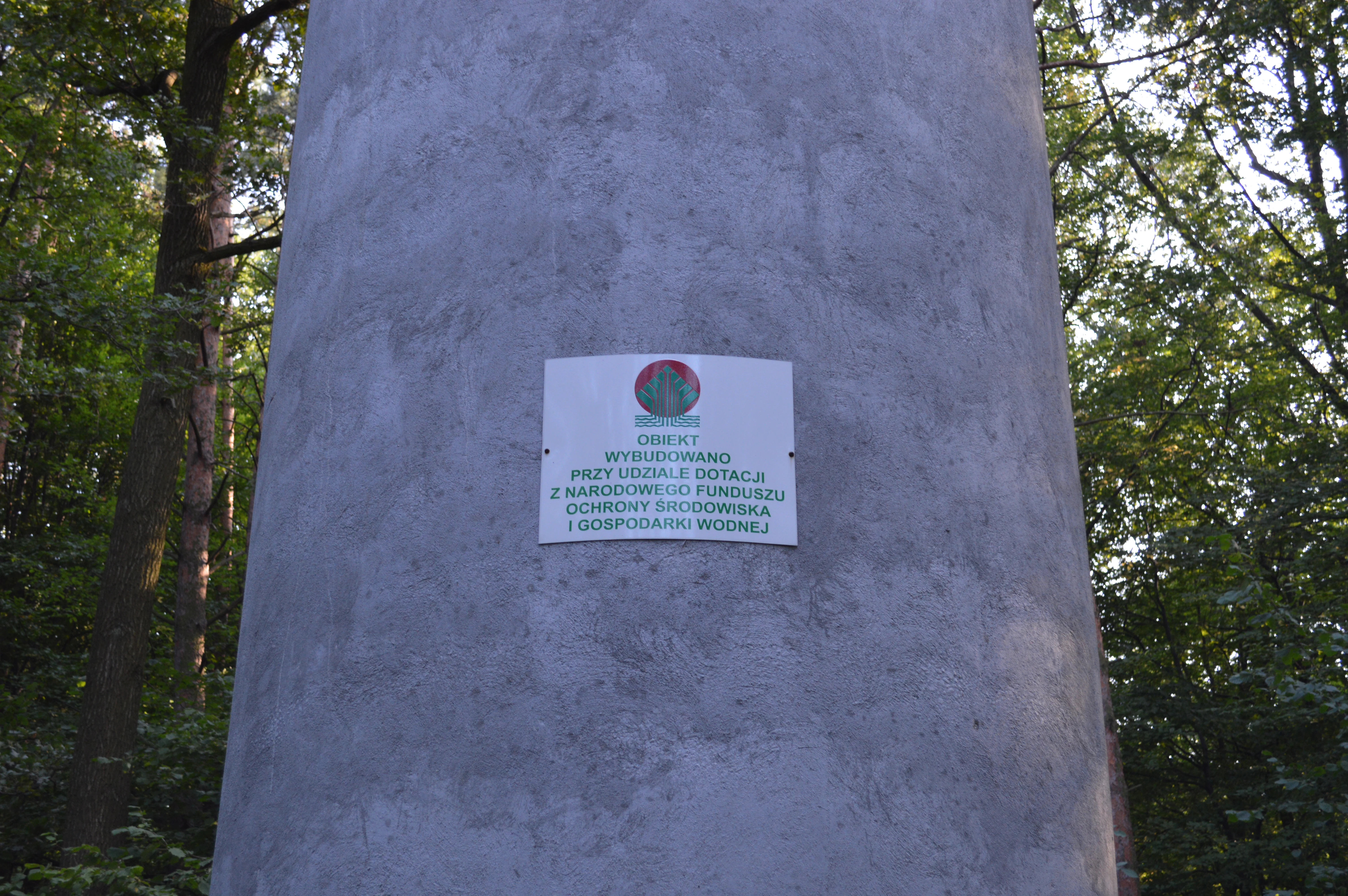A close-up view of a gray concrete surface featuring an informational sign with Polish text, mounted in a wooded area.