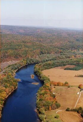 An aerial shot looking down at a winding dark blue river surrounded by dense forest showing autumn colors and open fields of brown grass.