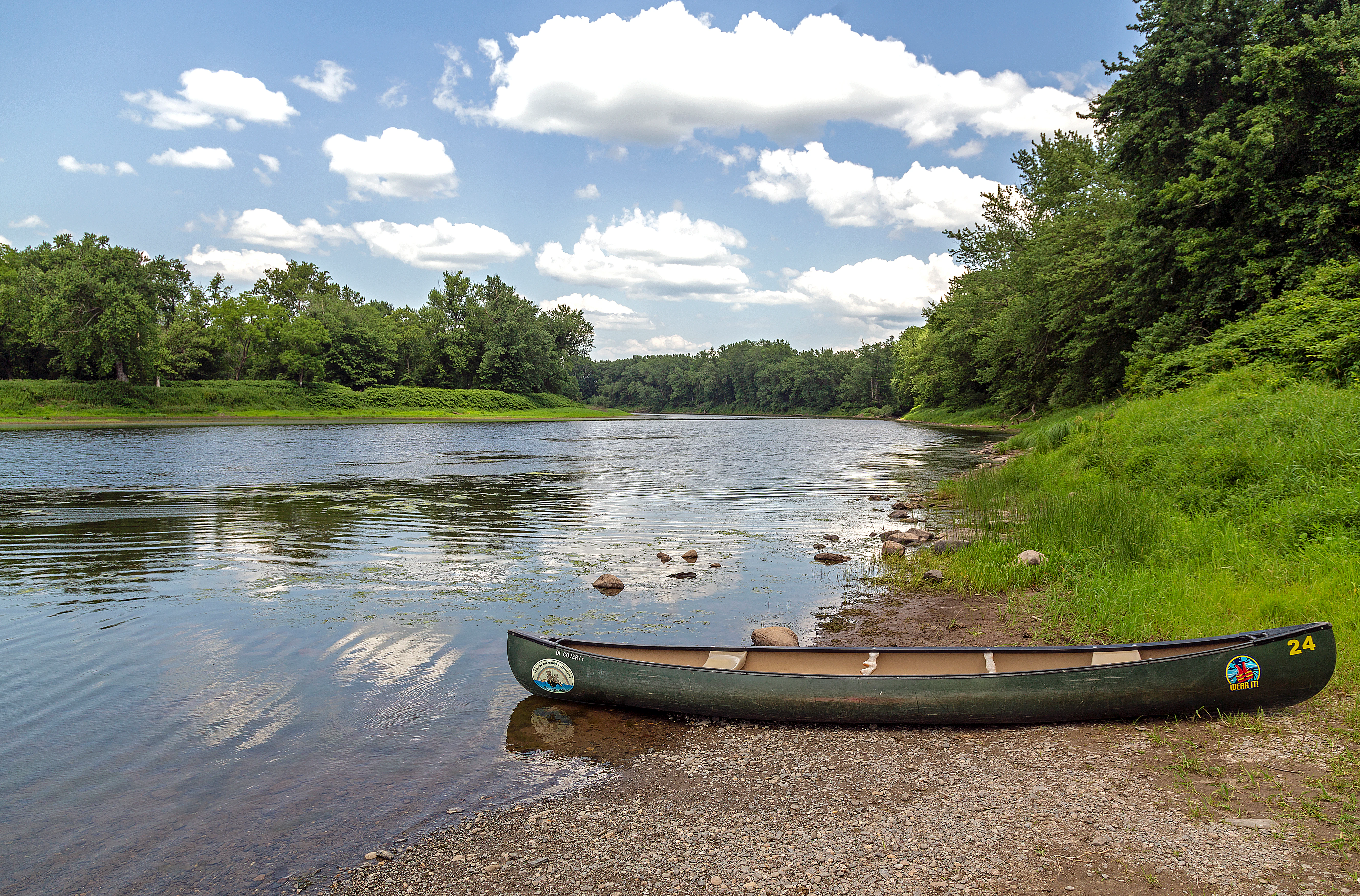 A green canoe pulled up onto a gravel riverbank with a wide, calm river and tree-lined banks in the background under a blue, partly cloudy sky.