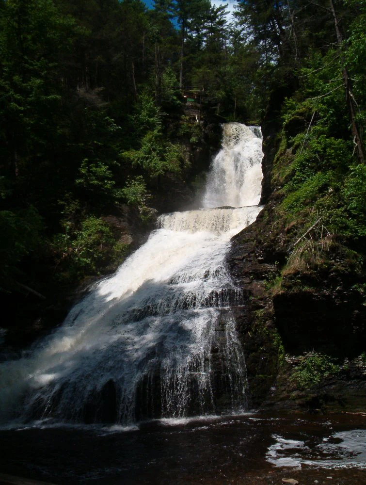 A multi-tiered waterfall cascading down a dark, forested rock cliff, surrounded by dense green trees.
