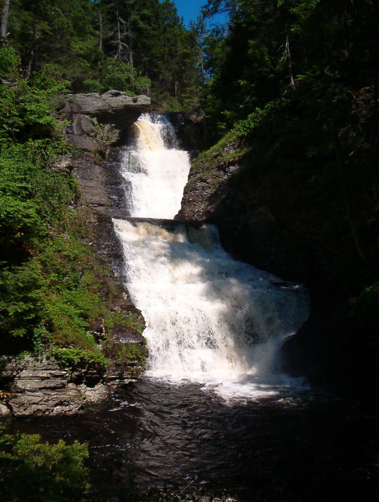 A multi-tiered waterfall cascading over dark rock ledges, surrounded by dense green forest foliage.