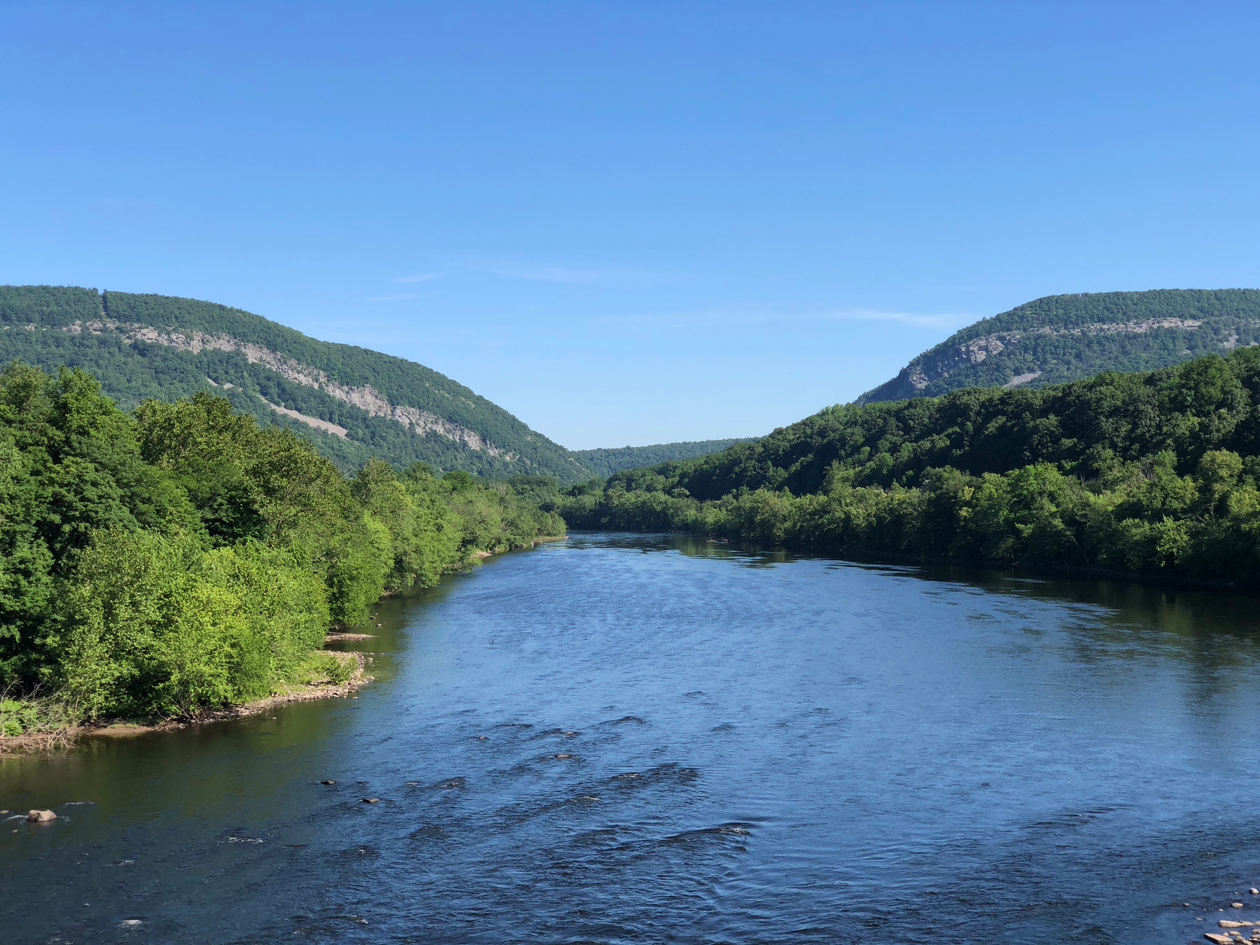 A wide view of the Delaware River flowing between two steep, forested mountains under a clear blue sky.