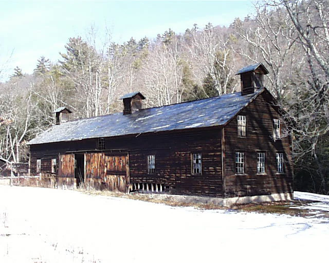 A weathered dark brown wooden barn with two roof cupolas sits in a snow-covered field against a backdrop of bare forest trees.