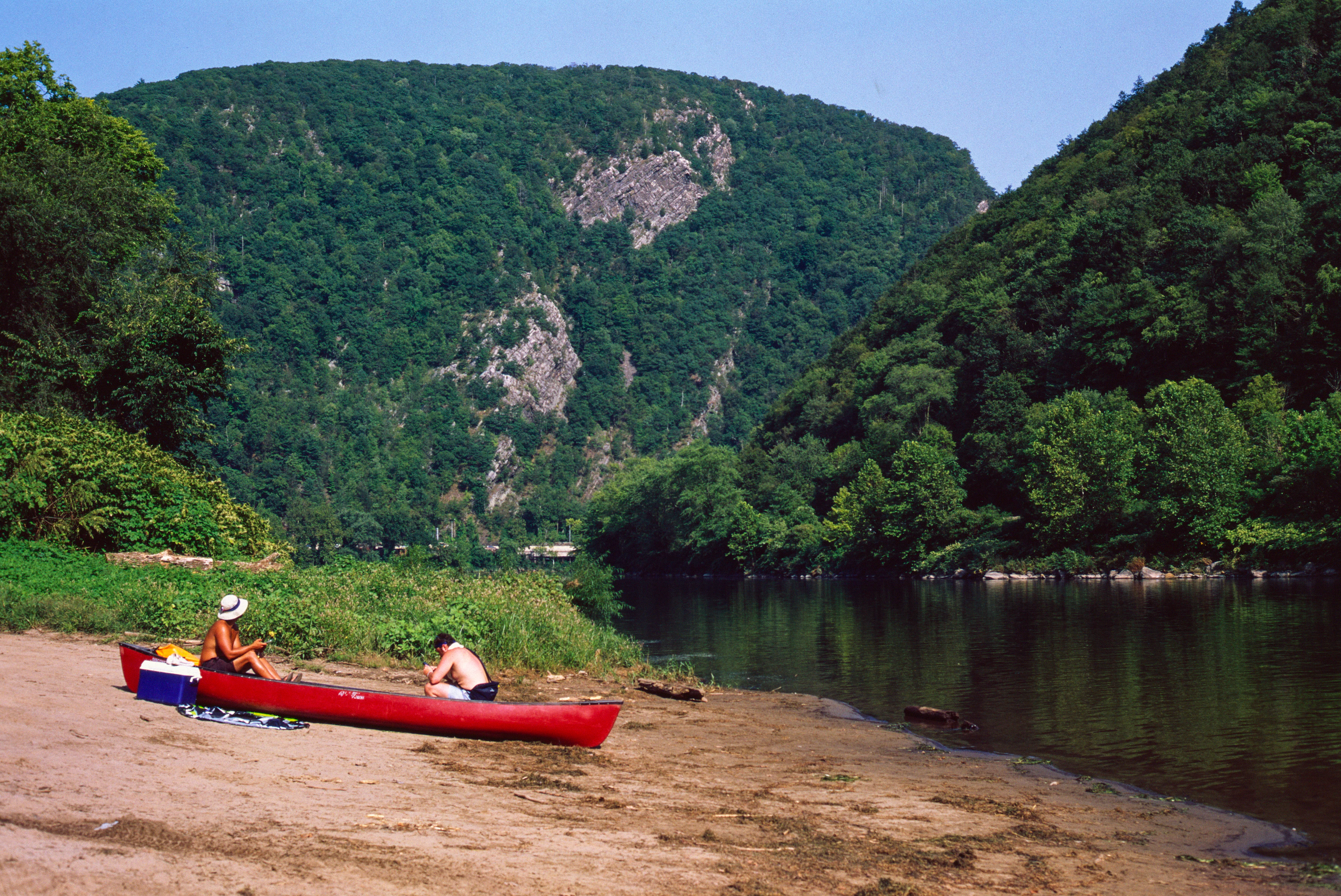 A wide shot showing two people sitting near a red canoe on a sandy riverbank, with steep, tree-covered mountains framing a river in the background.