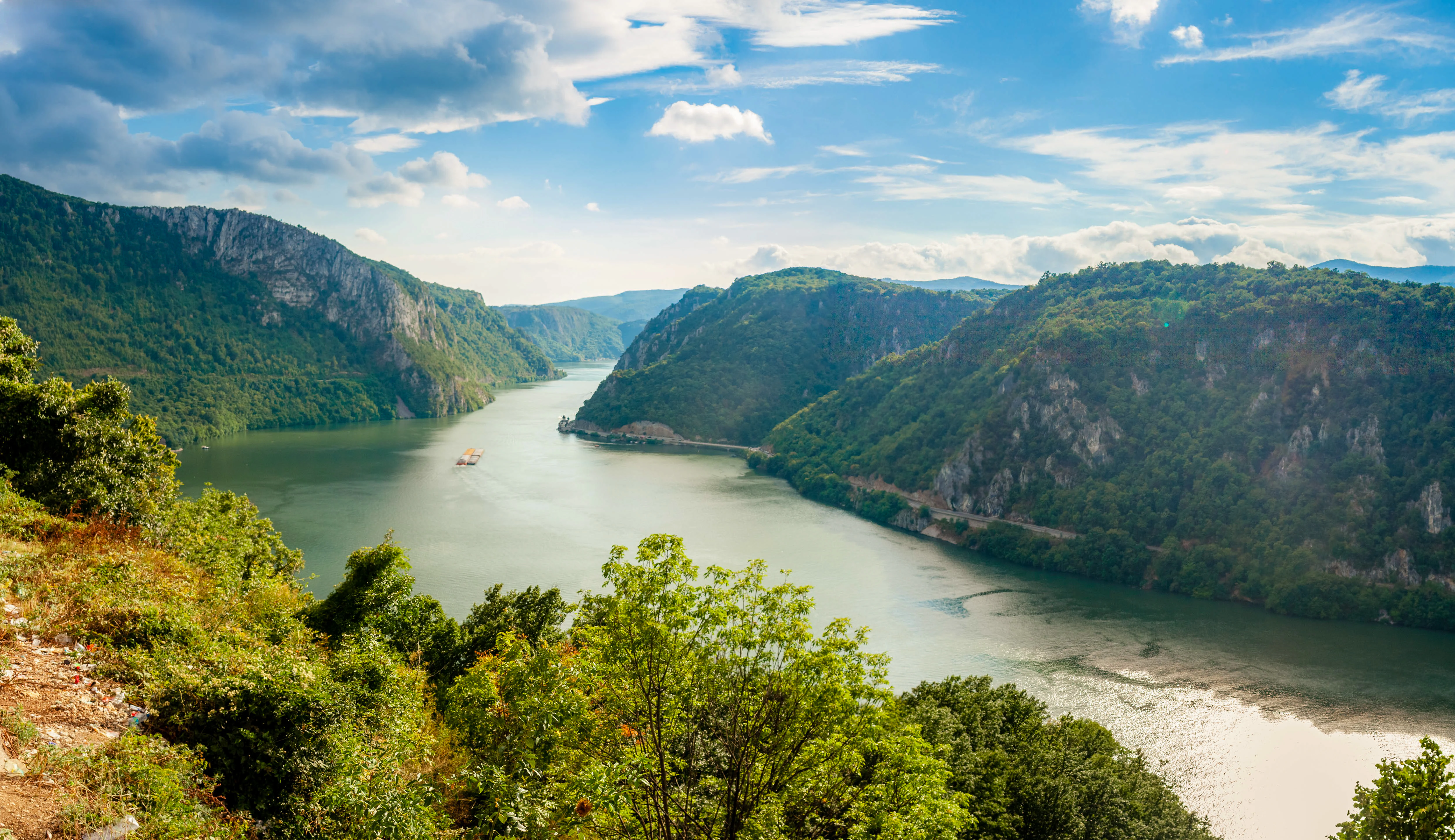 A wide view of the Danube River flowing through a narrow gorge between steep, forested mountains under a partly cloudy sky