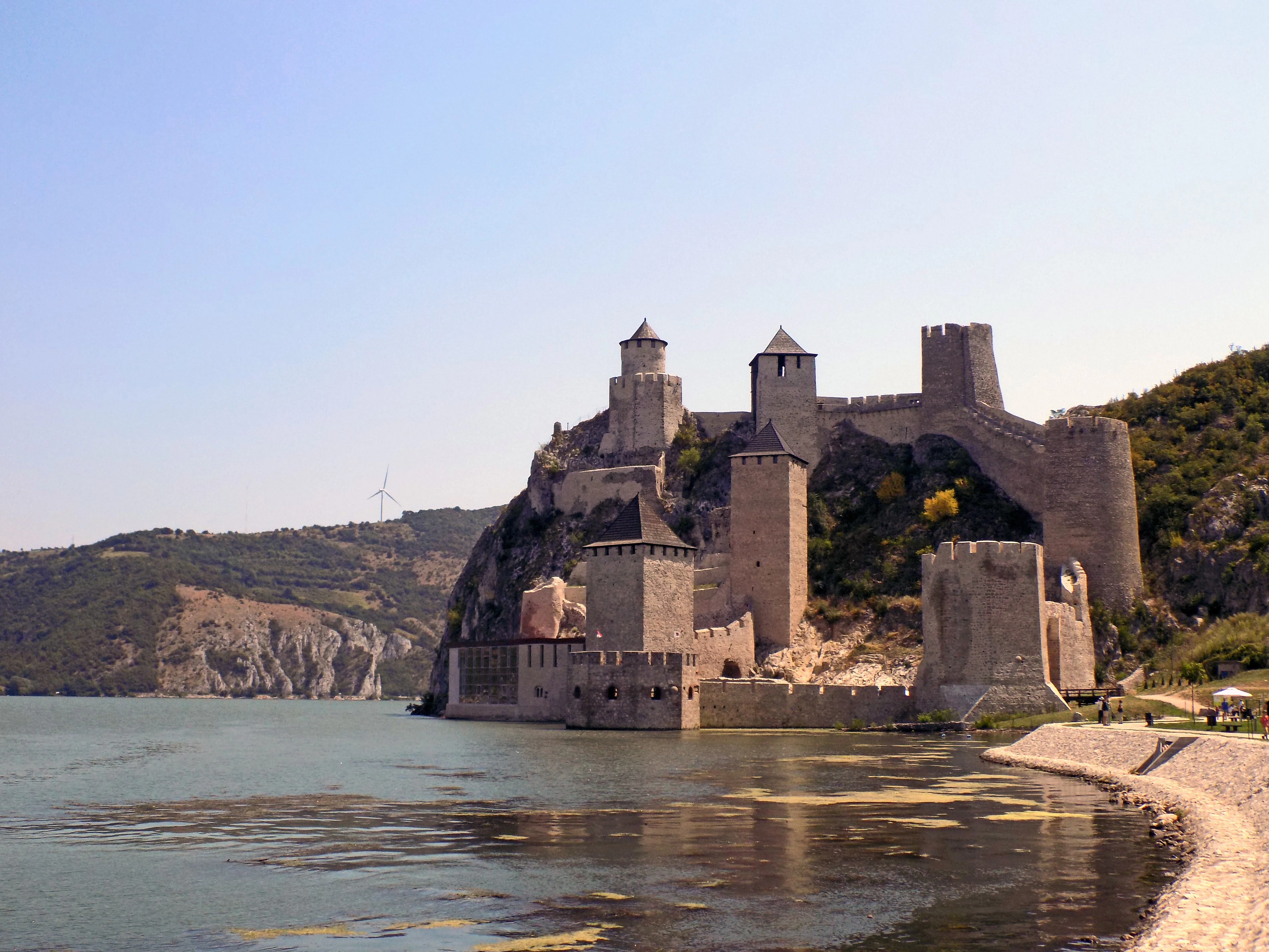 Stone fortress with multiple towers and walls on a riverbank, calm water, and hills under clear sky