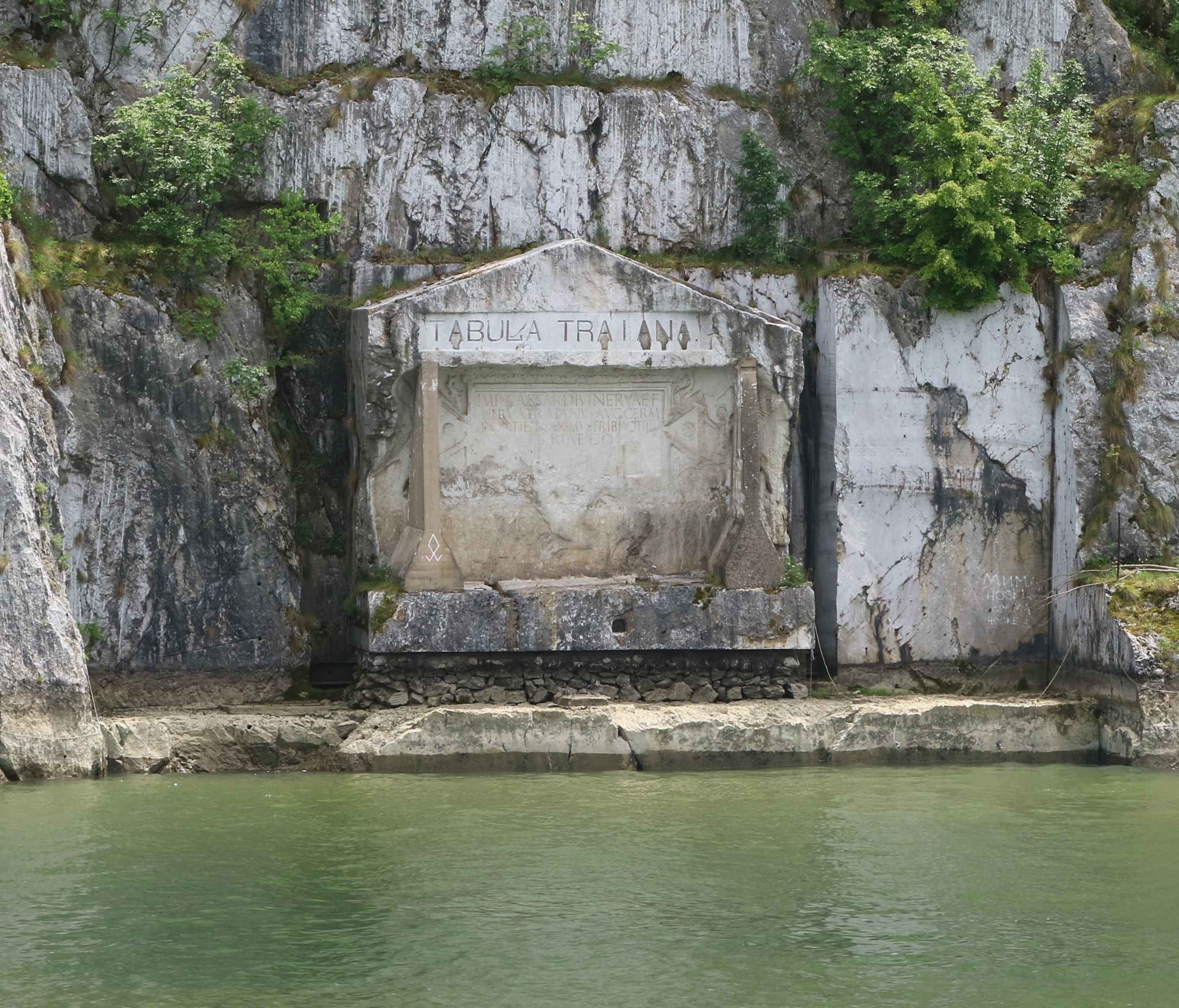 Stone monument with inscribed text 'TABULA TRAIANI' on a rocky cliffside above a body of water