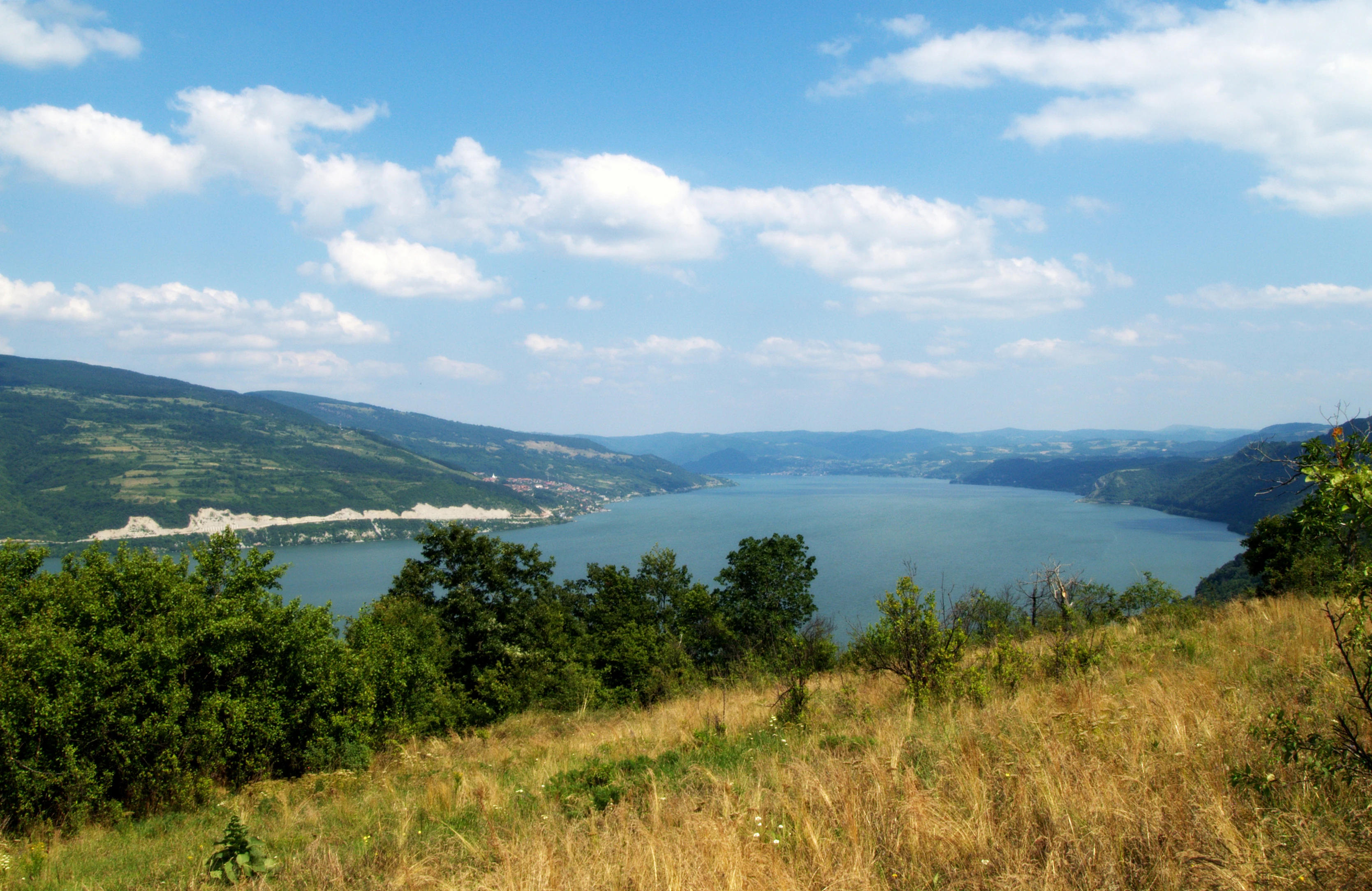A landscape view of a wide river, hilly terrain, and green vegetation under a blue sky with scattered clouds.