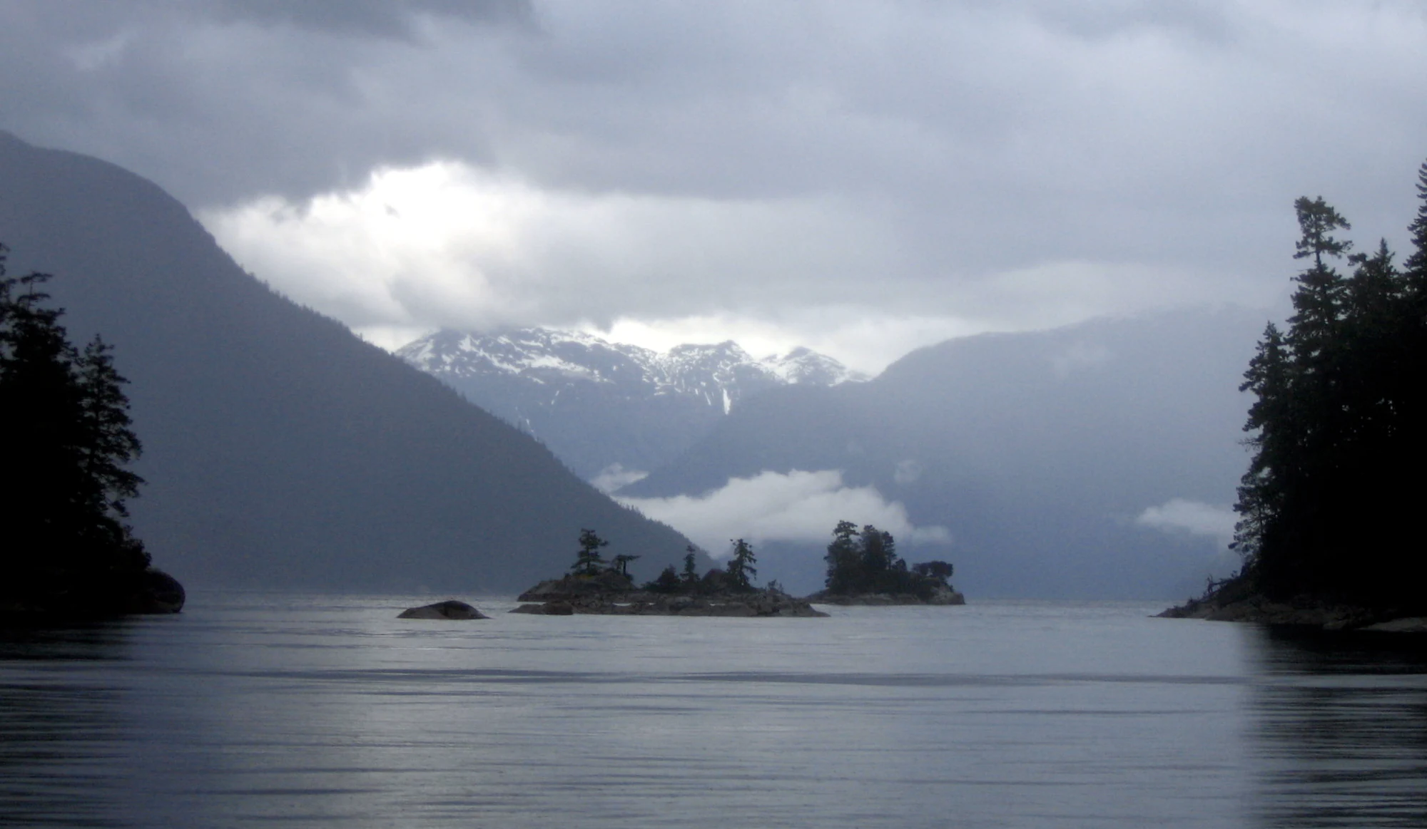 A wide, eye-level shot of a calm body of water framed by dense, dark evergreen forests on the left and right. Small, tree-covered rocky islands sit in the midground, with misty, snow-capped mountains rising in the background beneath heavy, overcast grey clouds.