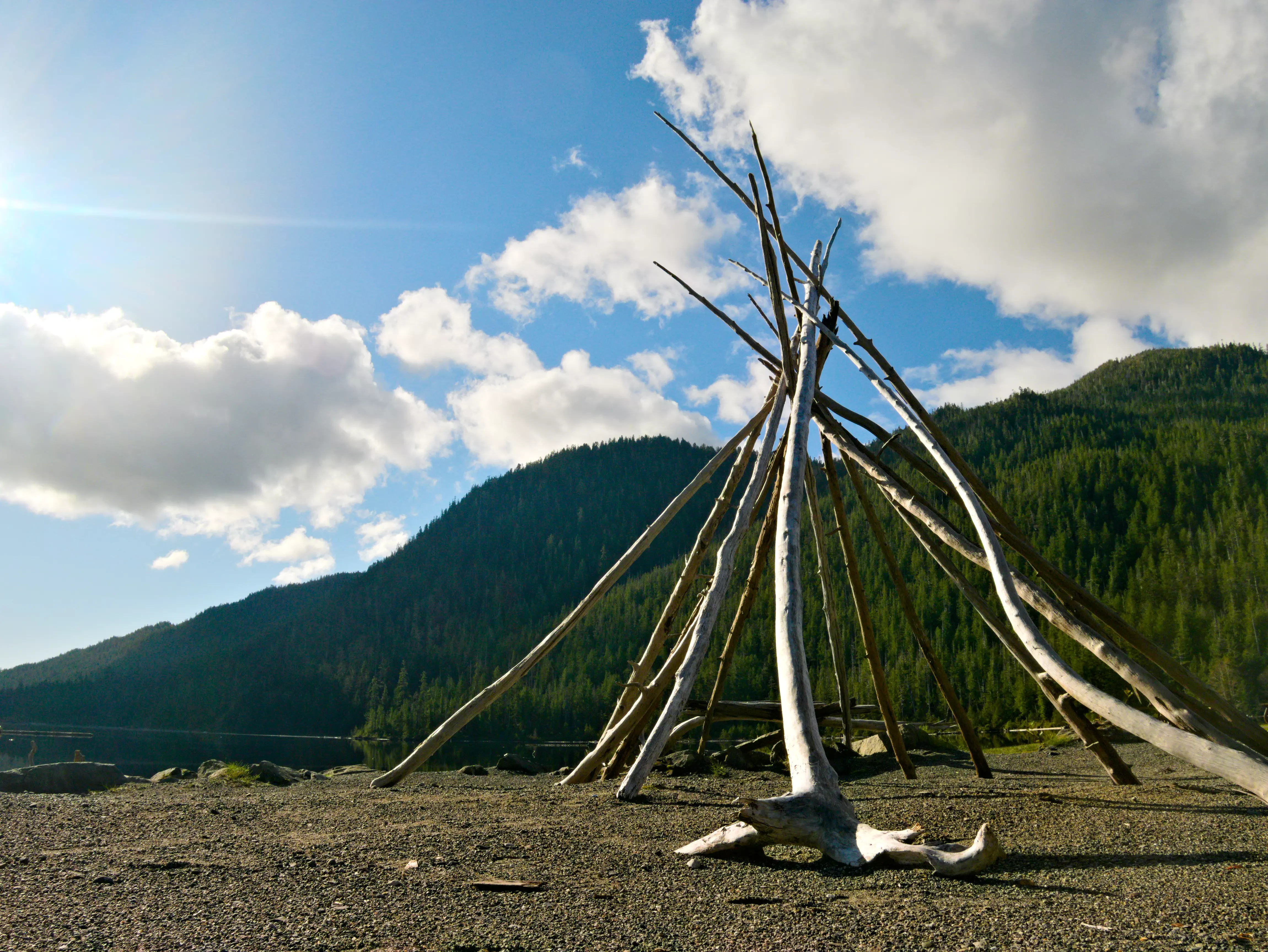 A teepee-shaped sculpture made of light-colored driftwood poles standing on a sandy lakeshore with a green, tree-covered mountain in the background under a blue sky with white clouds.