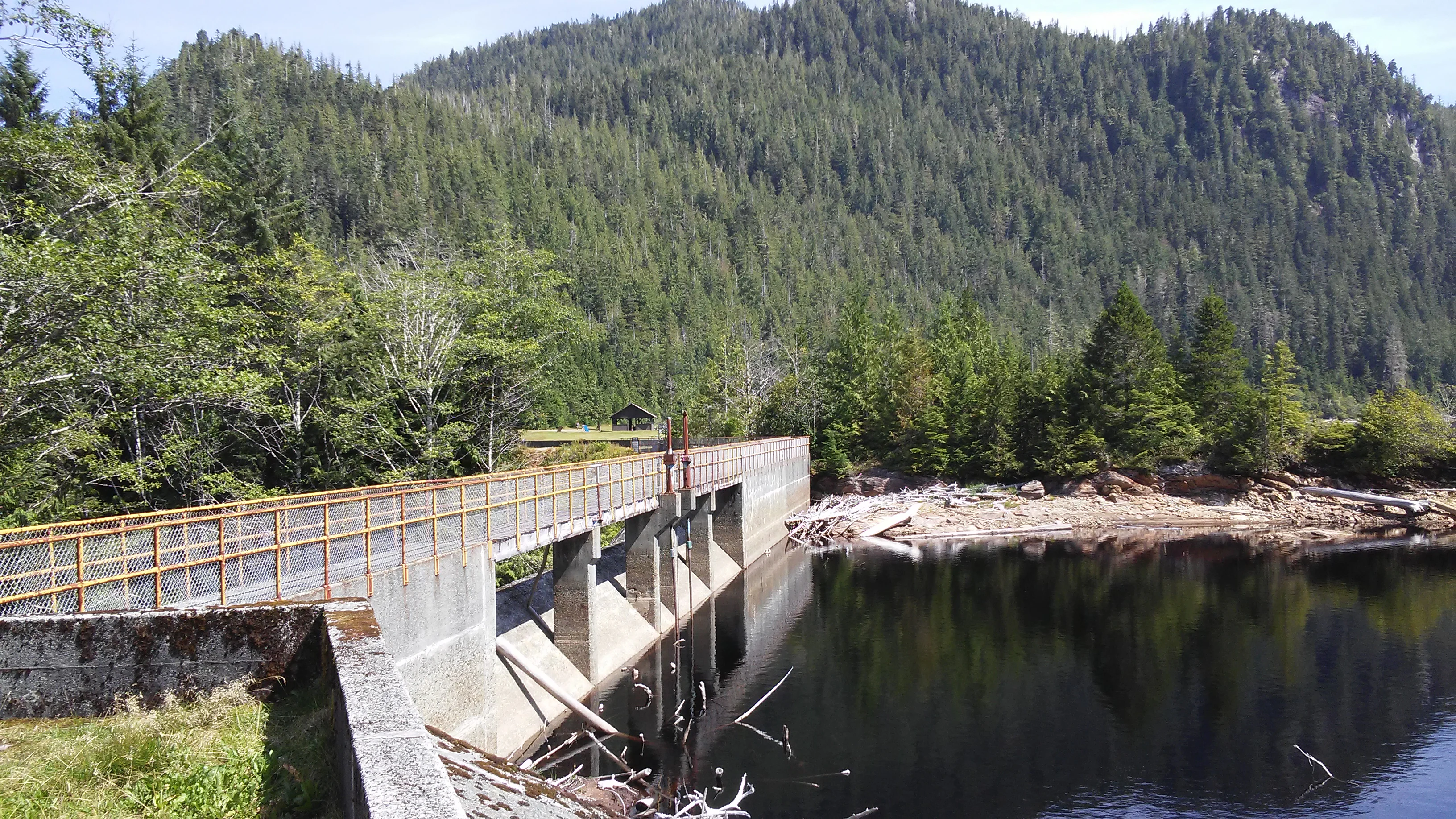 A concrete dam with a metal railing crossing a section of Diana Lake, backed by a densely forested mountainside.