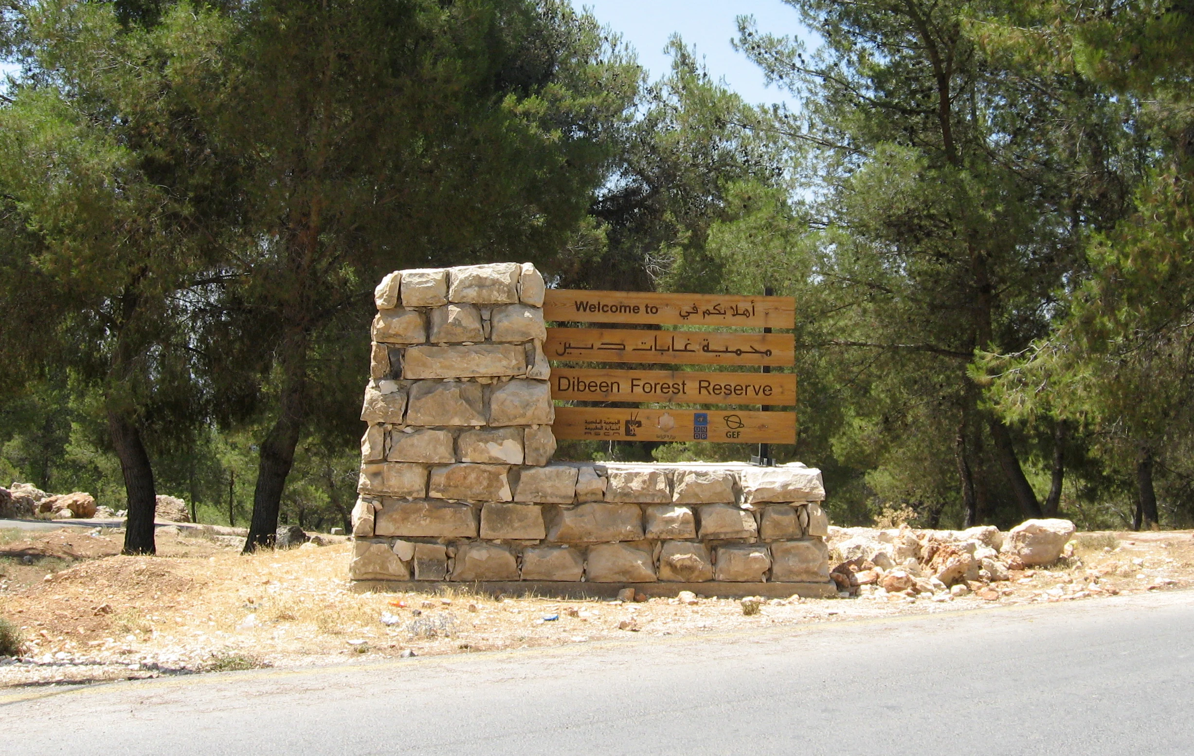 Estructura de piedra con un letrero de madera que muestra el texto 'Dibeen Forest Reserve', rodeada de árboles y una carretera