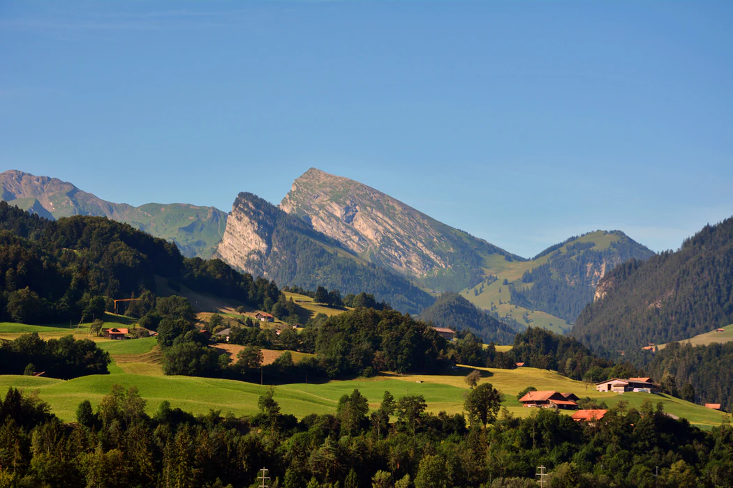 Una amplia toma panorámica a nivel de los ojos de un valle montañoso verde con colinas onduladas, parches de bosque denso y granjas tradicionales de madera bajo un cielo azul despejado.