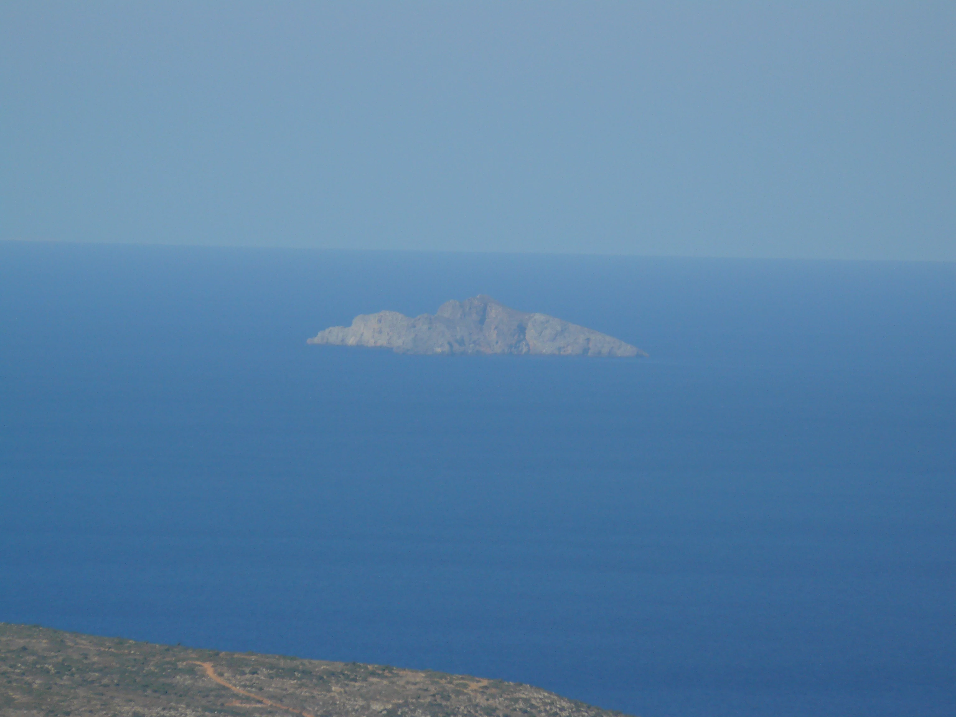 A small, rocky, uninhabited island sits in the middle of a calm, blue Mediterranean sea, viewed from a high vantage point.