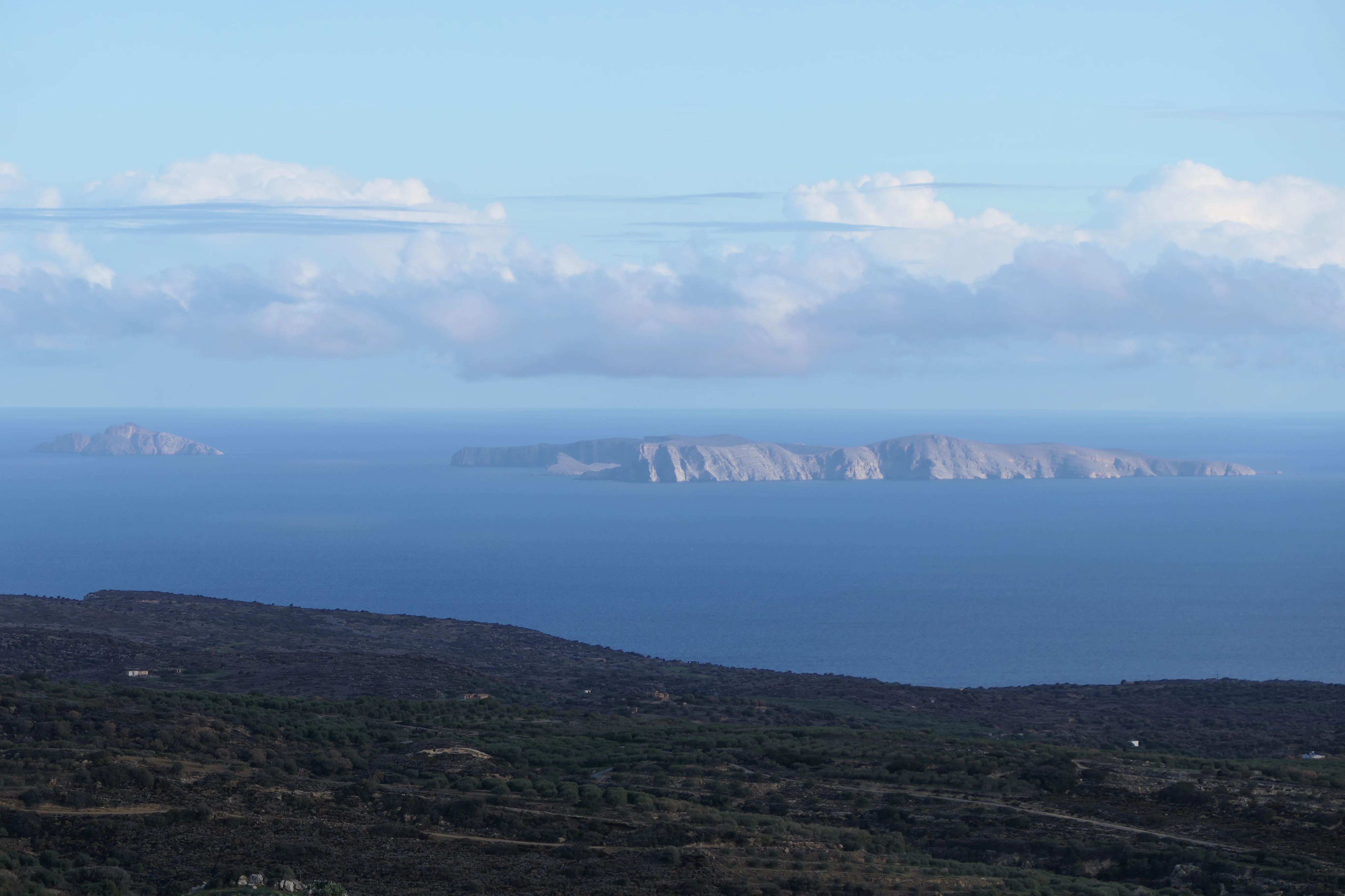 A distant view of the rocky Dionysades islands rising above the blue Aegean Sea, seen from the scrub-covered slopes of the Cretan coast.