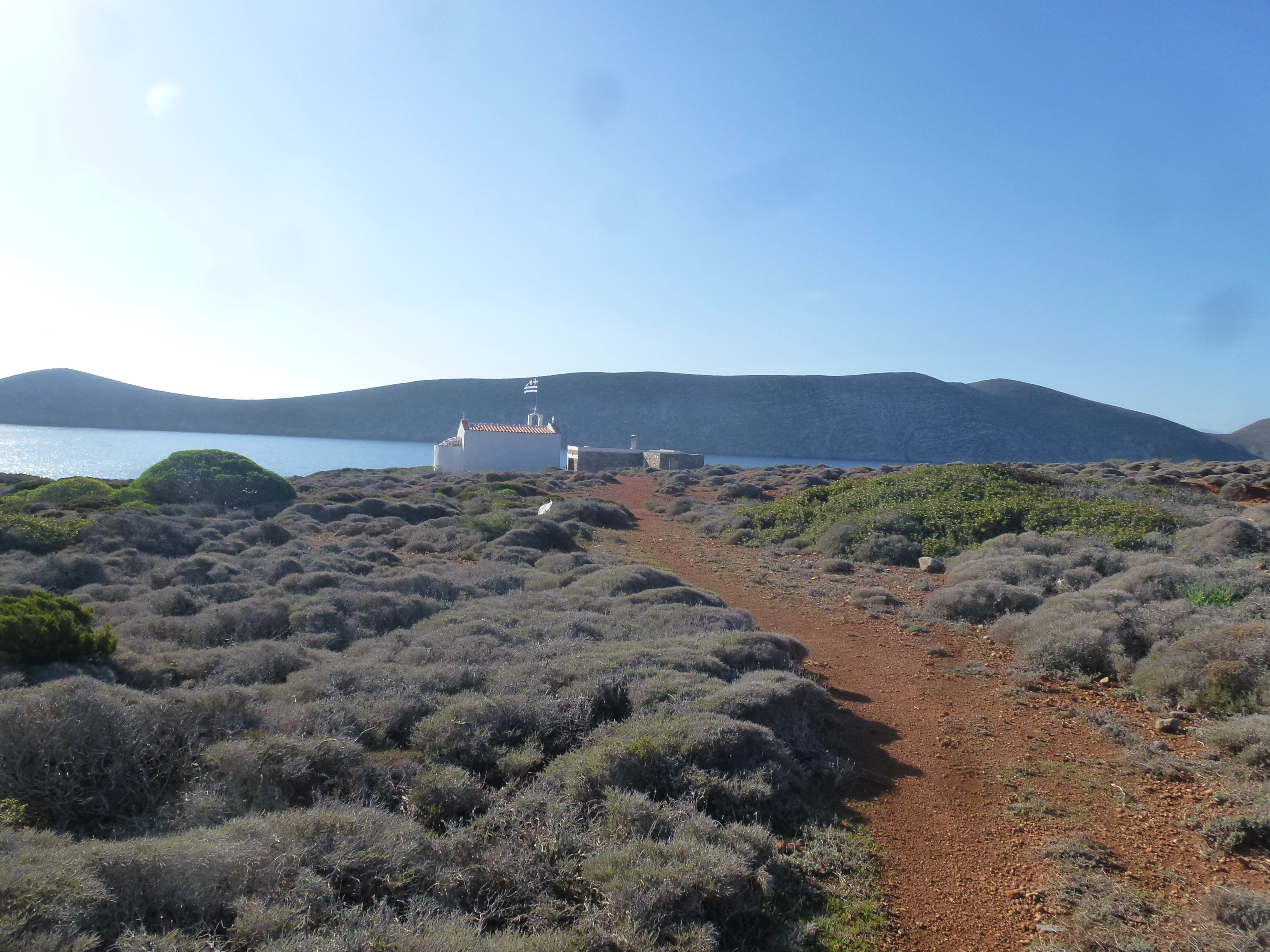 A red dirt path winds through low lying scrub brush toward a small white chapel on a coastline with a hilly island in the background.