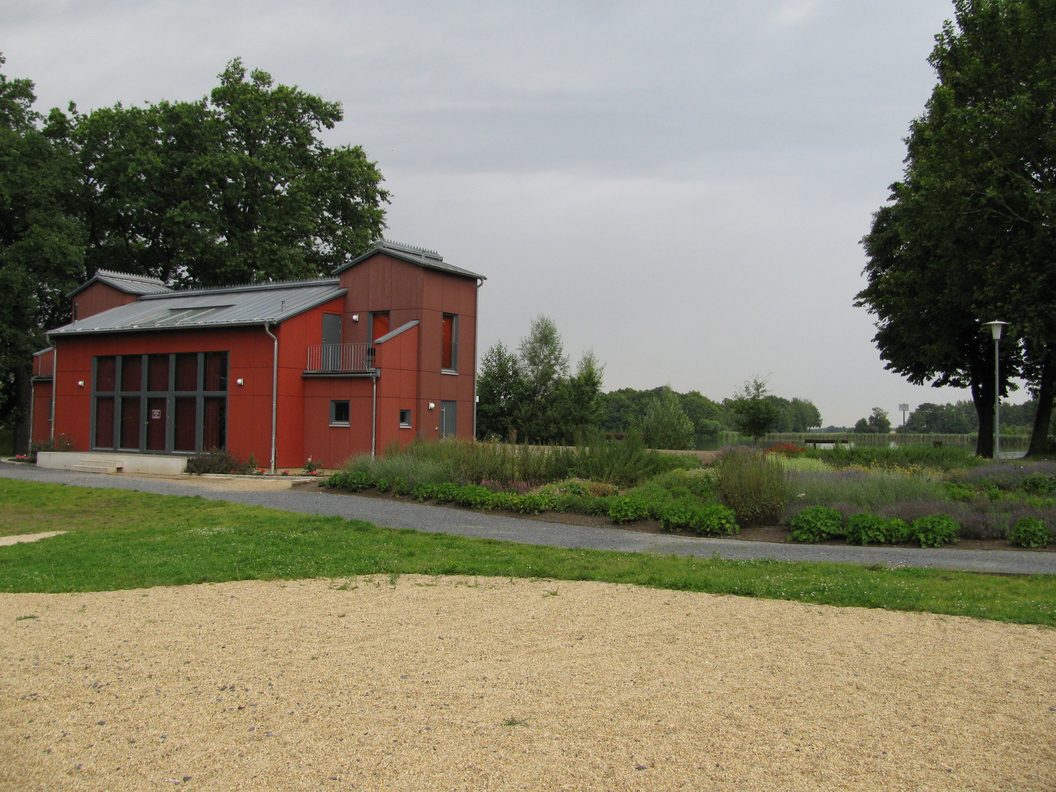 A modern two story red building with a gray metal roof stands next to a paved path, surrounded by grass and low shrubs under an overcast sky.