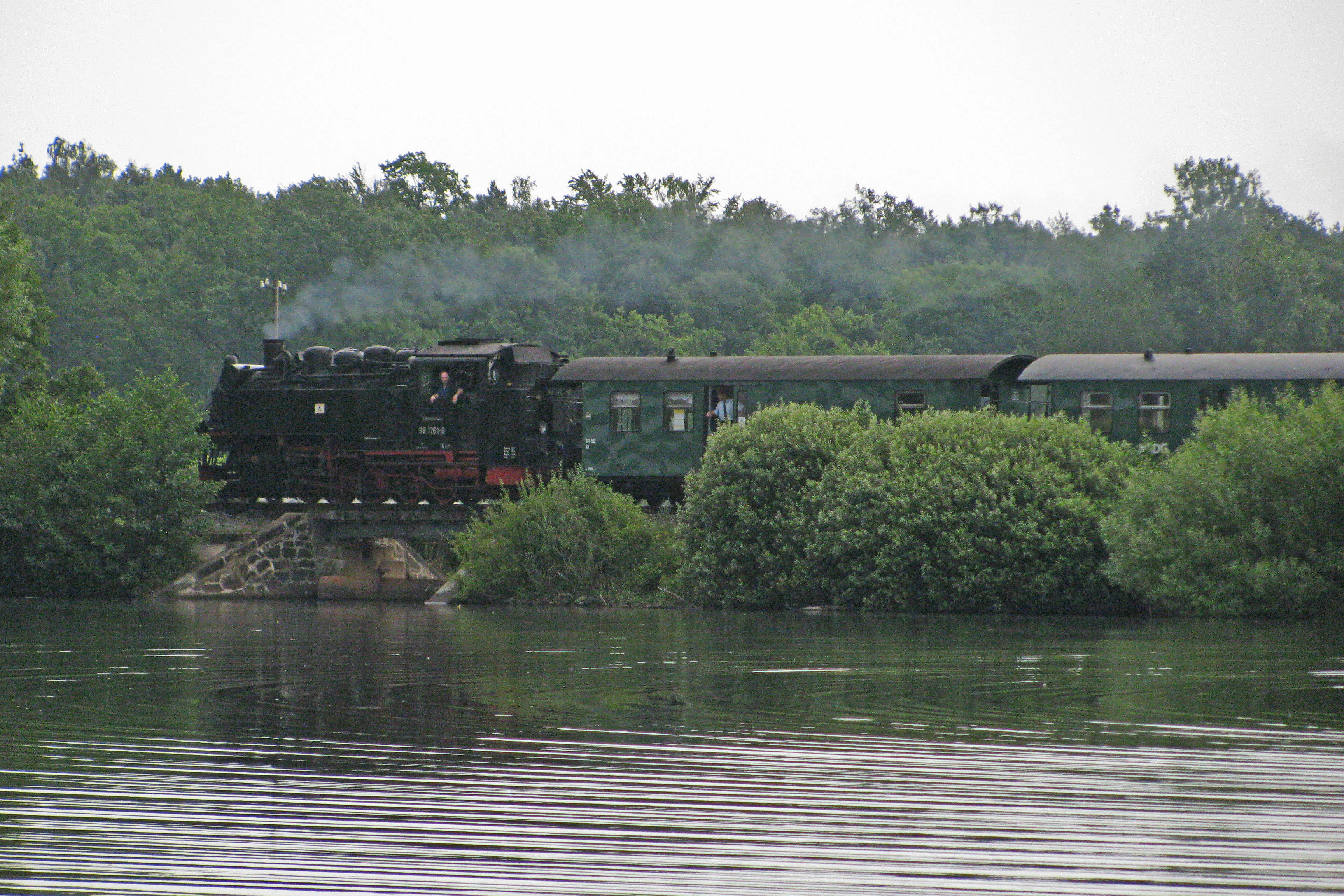 A vintage steam locomotive pulling passenger carriages across a bridge over a lake, with dense forest in the background.