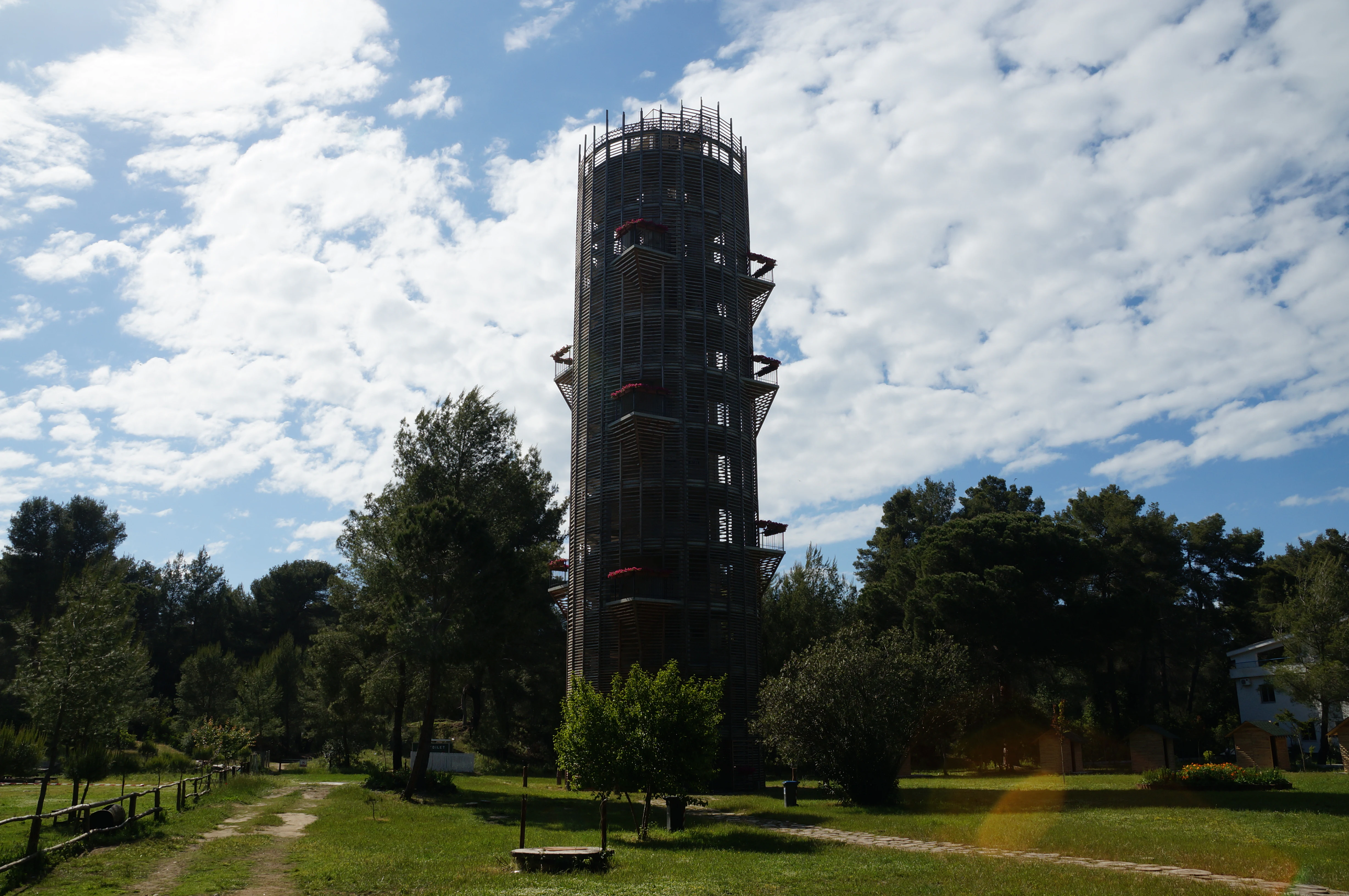 A tall metal observation tower with multiple platforms stands on a grassy field surrounded by trees under a partly cloudy sky
