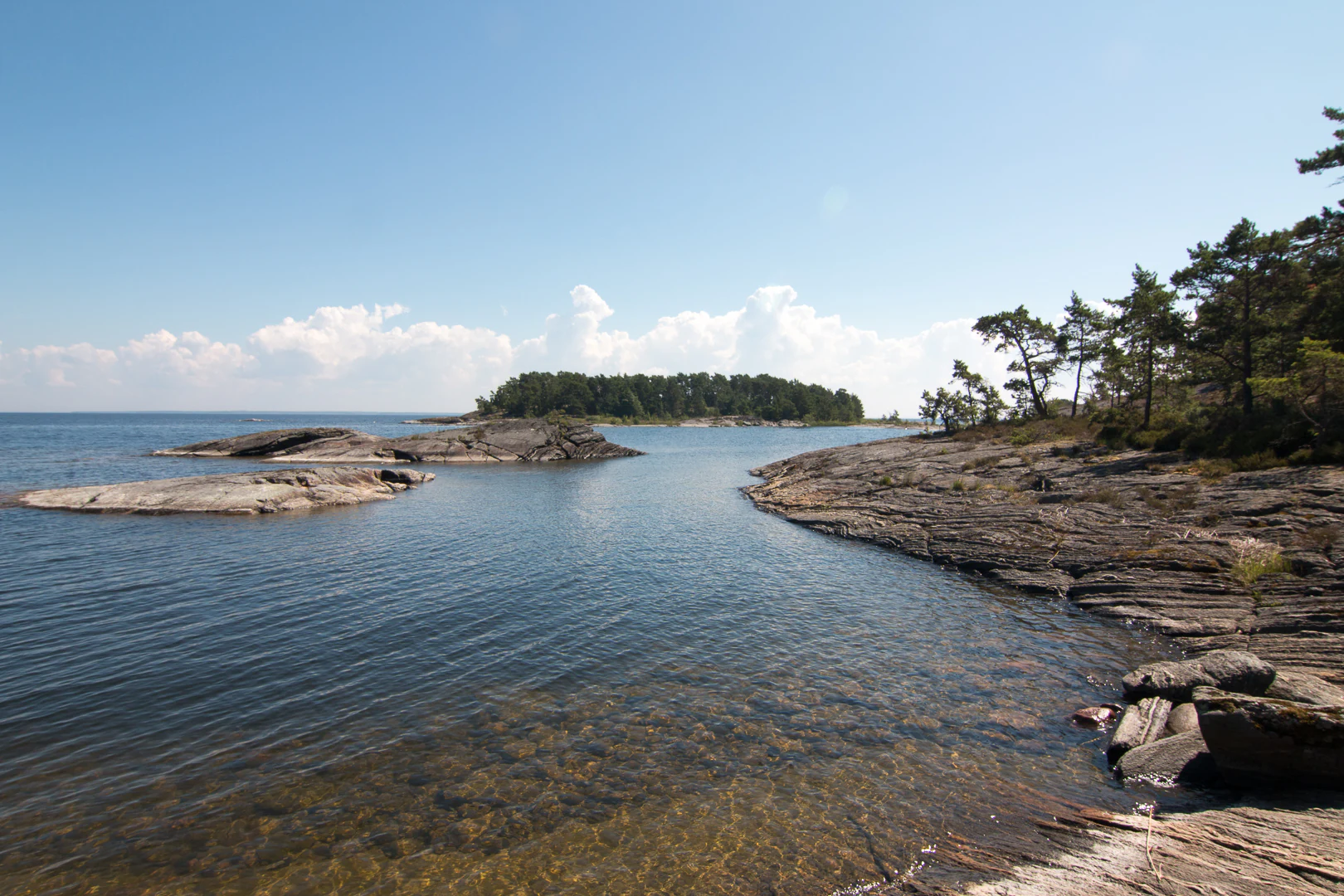 Rocky shoreline with clear water, distant islands, and trees on the right side under a clear blue sky